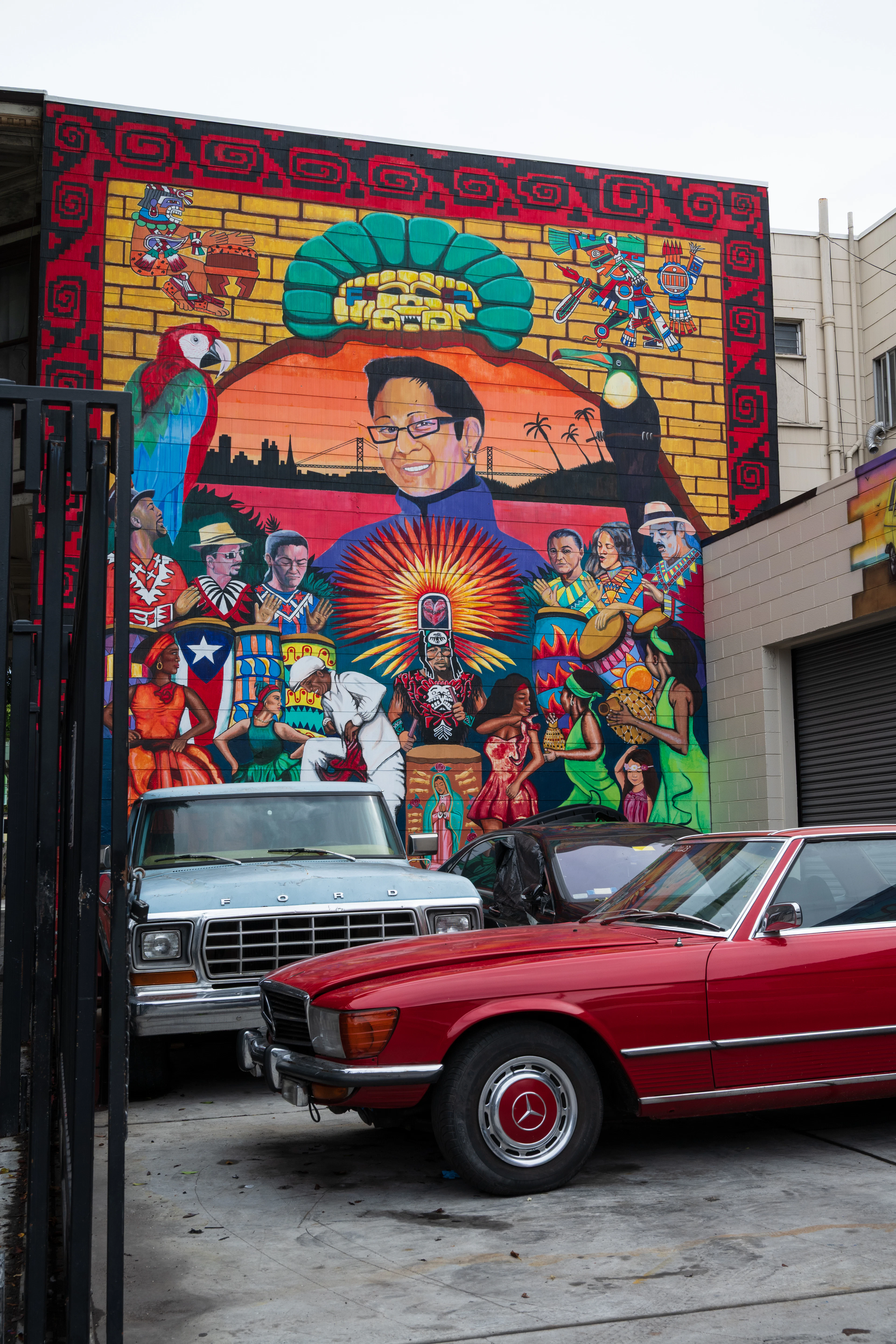 A large Mission District mural in San Francisco covering a whole wall — Aztec motifs, a scarlet macaw, dancers and musicians around a central portrait, with the city's bridges painted into the skyline behind — with two old cars parked in front of it