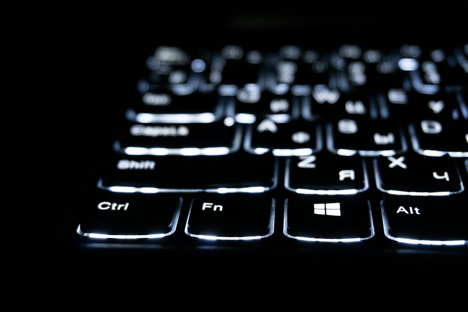 A backlit laptop keyboard glowing in a dark room, keys lit from beneath with the rest of the desk in shadow