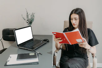 Woman reading a book at a desk with laptop.