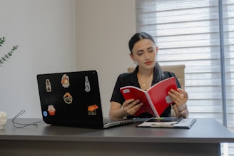 A woman reading a red book at a desk.