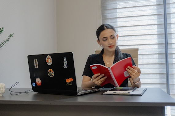 A woman reading a red book at a desk.