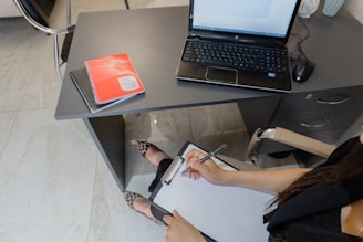 Person writing on clipboard at desk with laptop.