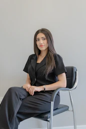 A woman in a black suit sits on a chair.