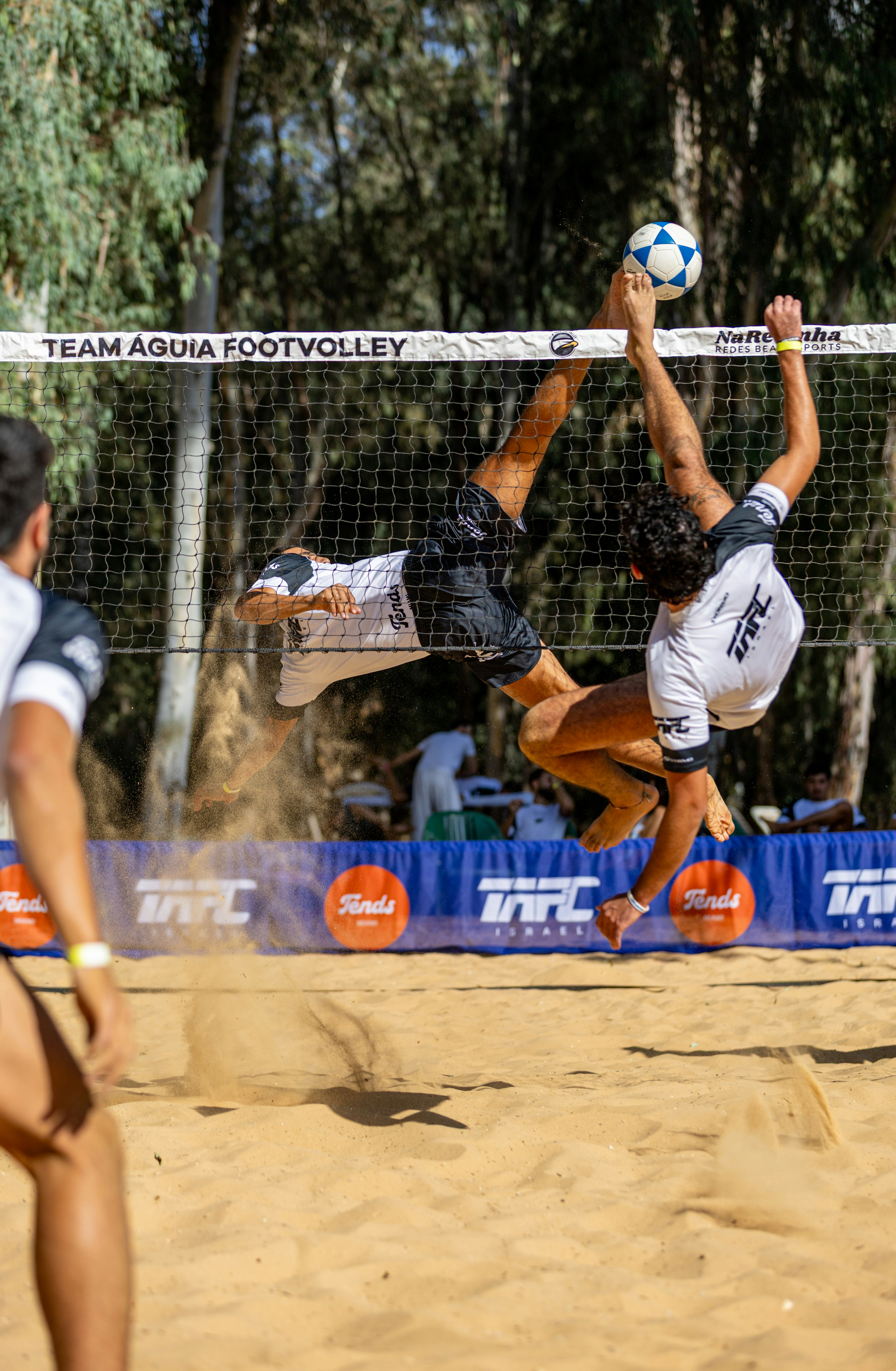 Dua pria bermain footvolley di lapangan berpasir.