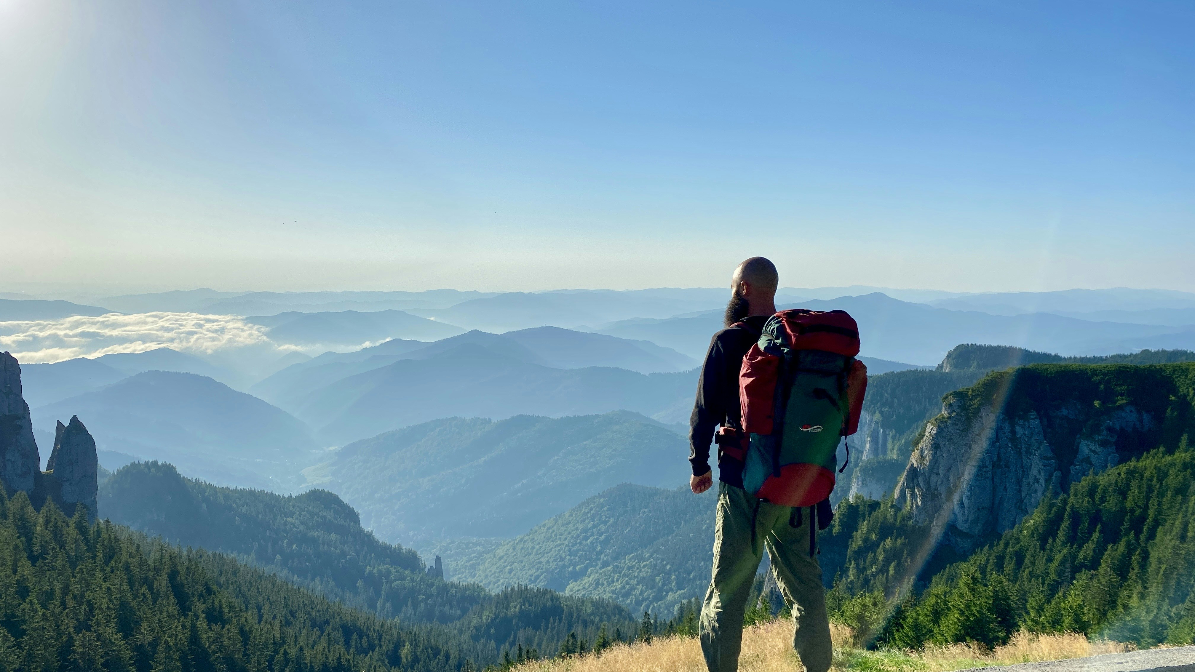 A couple wearing backpacks stands on a scenic overlook, gazing at a vast mountain range, representing the freedom and adventure achieved after selling their home.