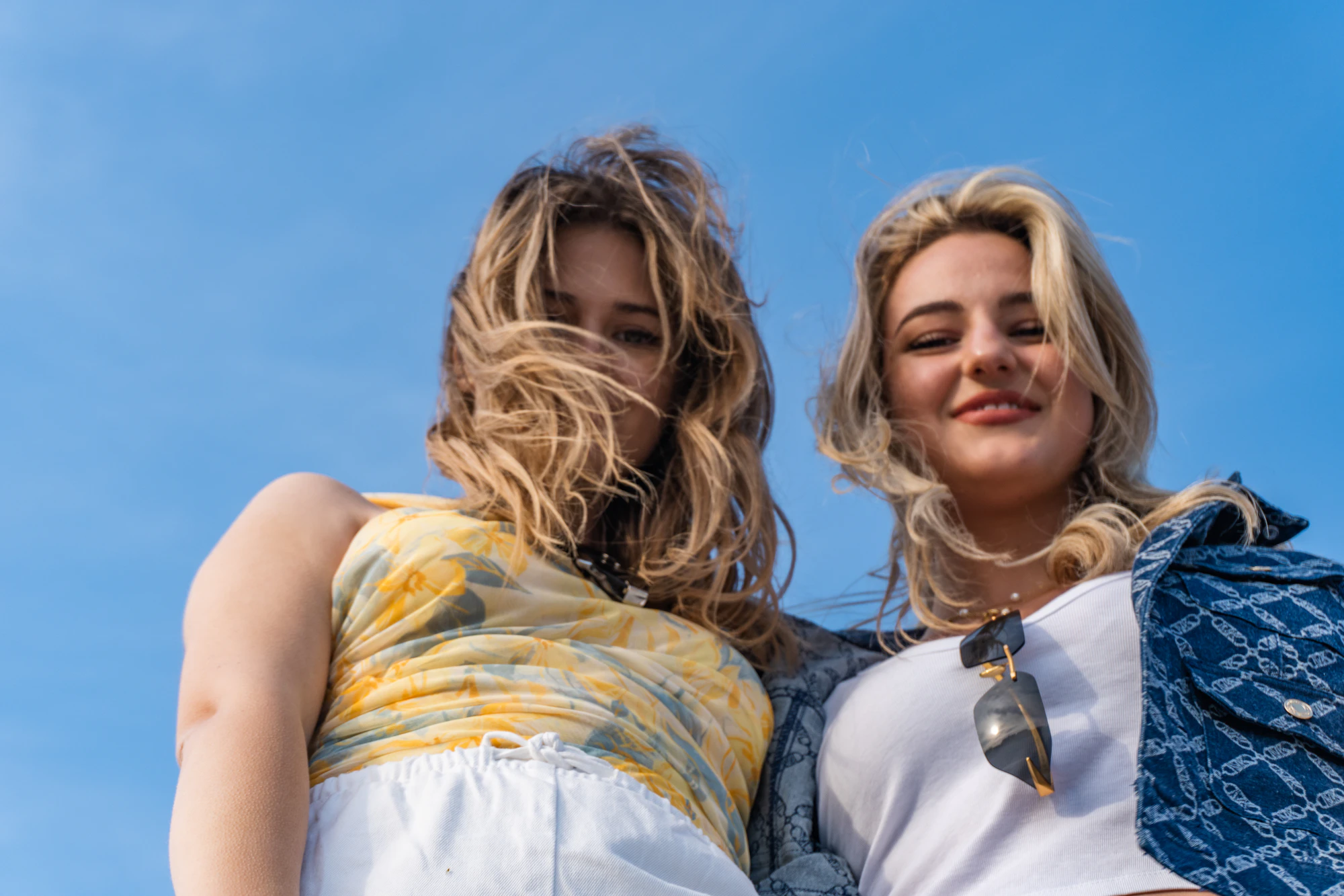 Two young women smiling down at the camera against a clear blue sky