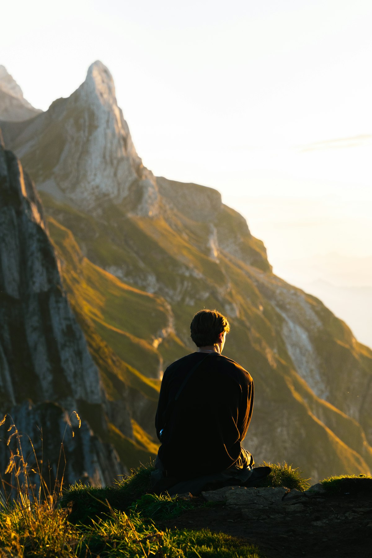 A person sitting on a grassy ledge, looking up at a sunlit mountain peak