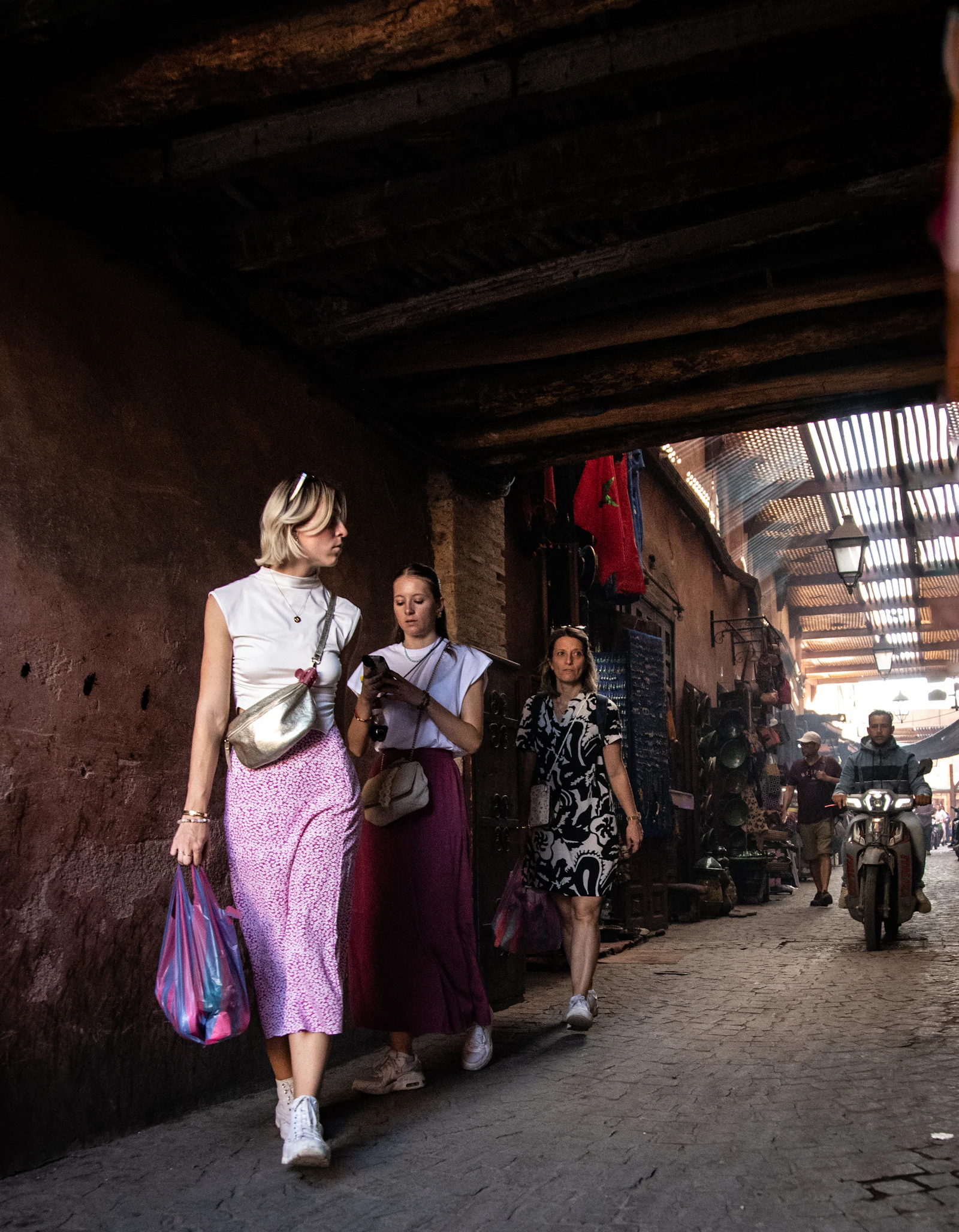 Visitors walking through a covered souk passage in the Marrakech medina, lined with shops and a passing scooter