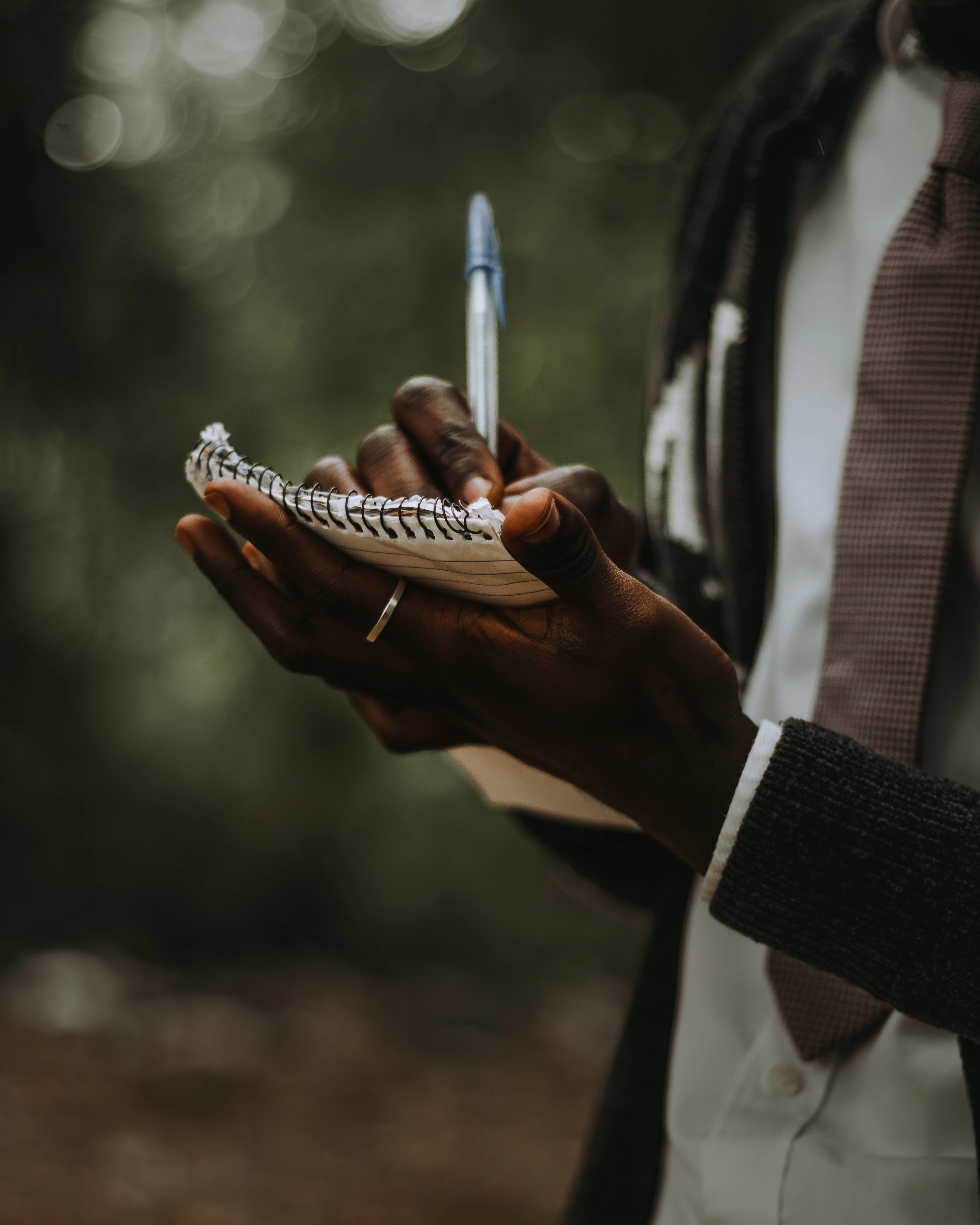 A person's hands writing in a small notebook resting on their knee, outdoors