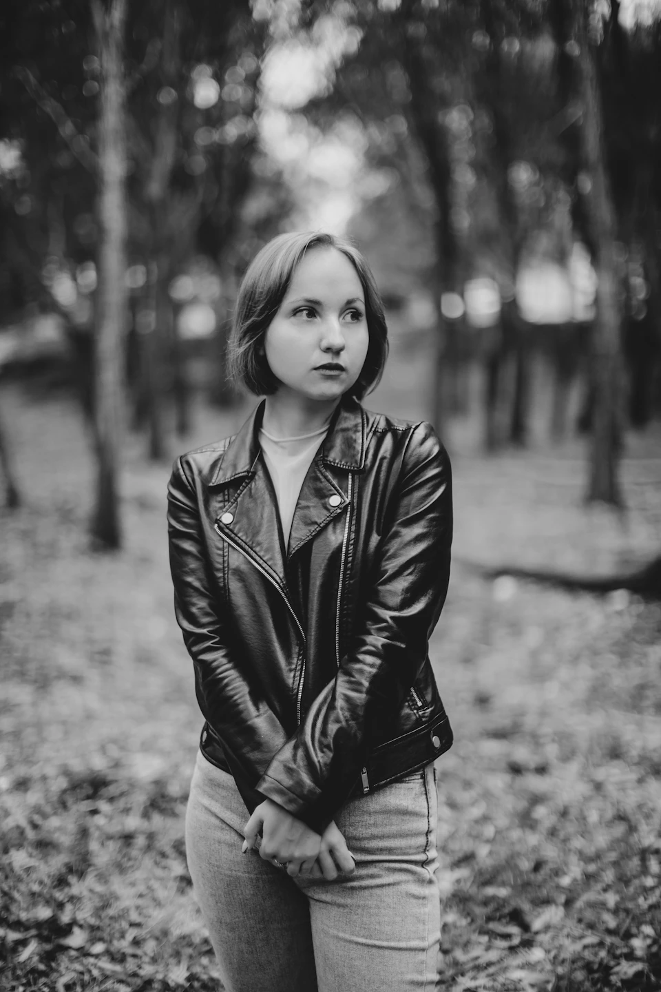 Young woman stands alone in forest wearing black leather jacket and jeans