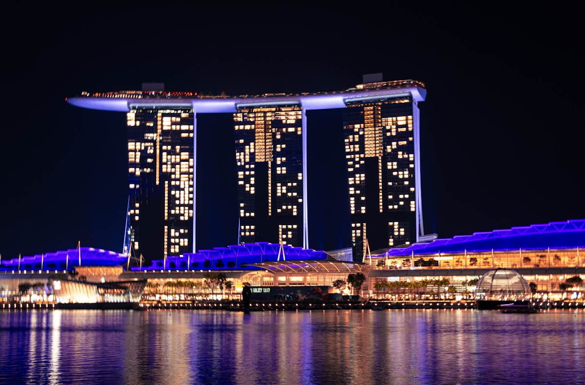 Marina Bay Sands hotel glows at night with its distinctive boat-shaped top deck, its reflection mirrored in the dark water below, surrounded by other illuminated city buildings.. Photo by Julius Hildebrandt on Unsplash.