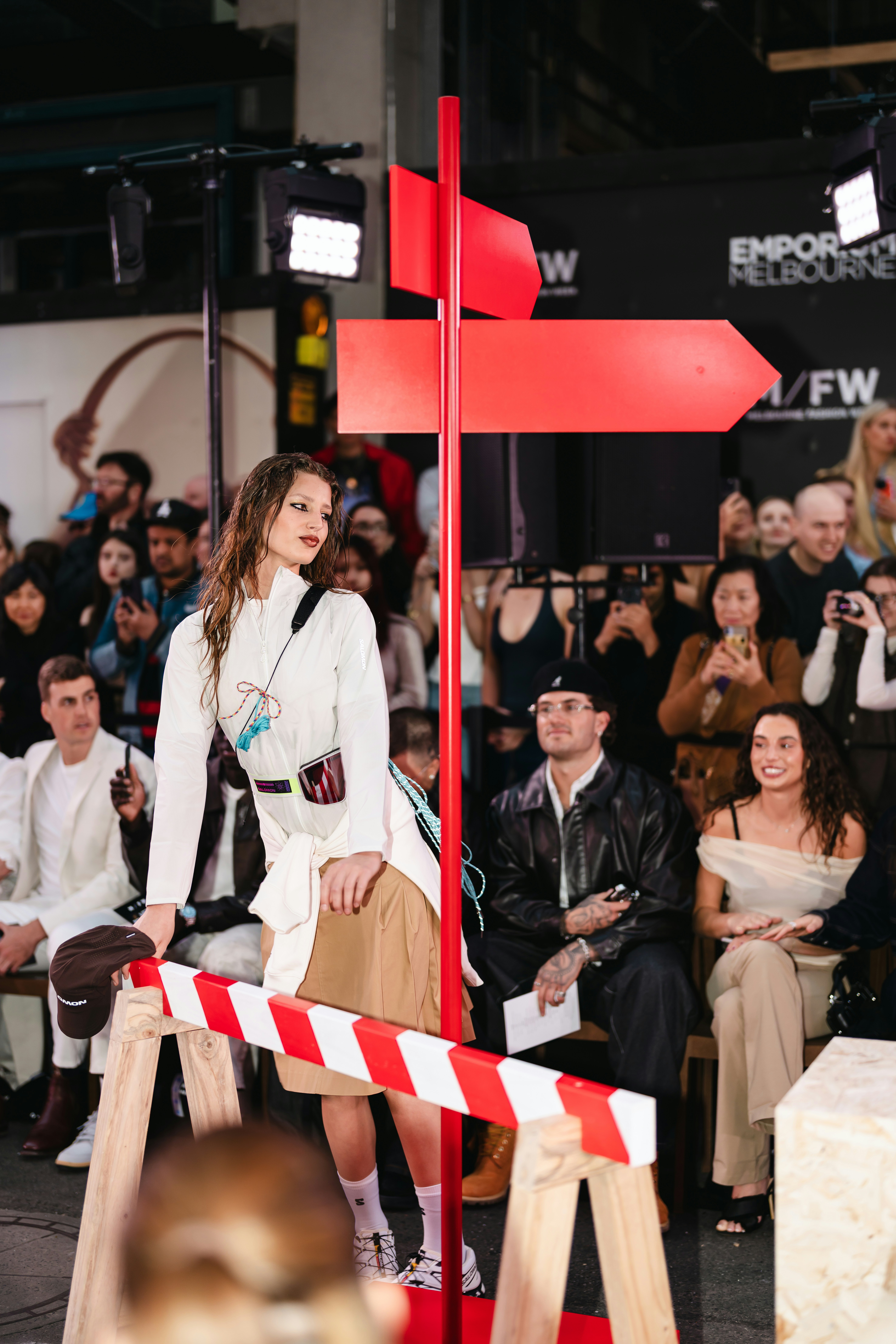 Model walks runway with red signpost and audience watching audience