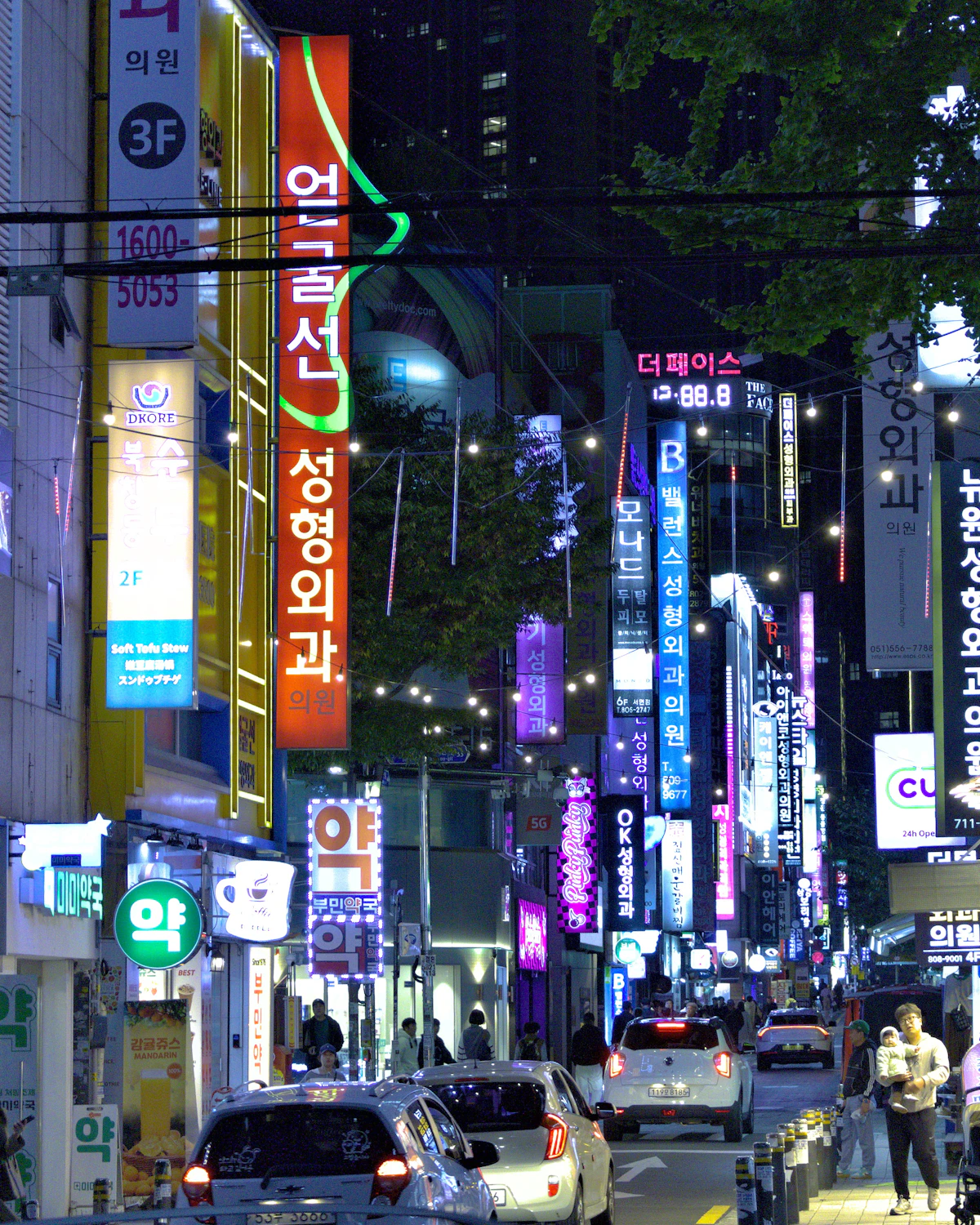 Narrow street at night packed with vertical neon signs in Korean, parked cars below
