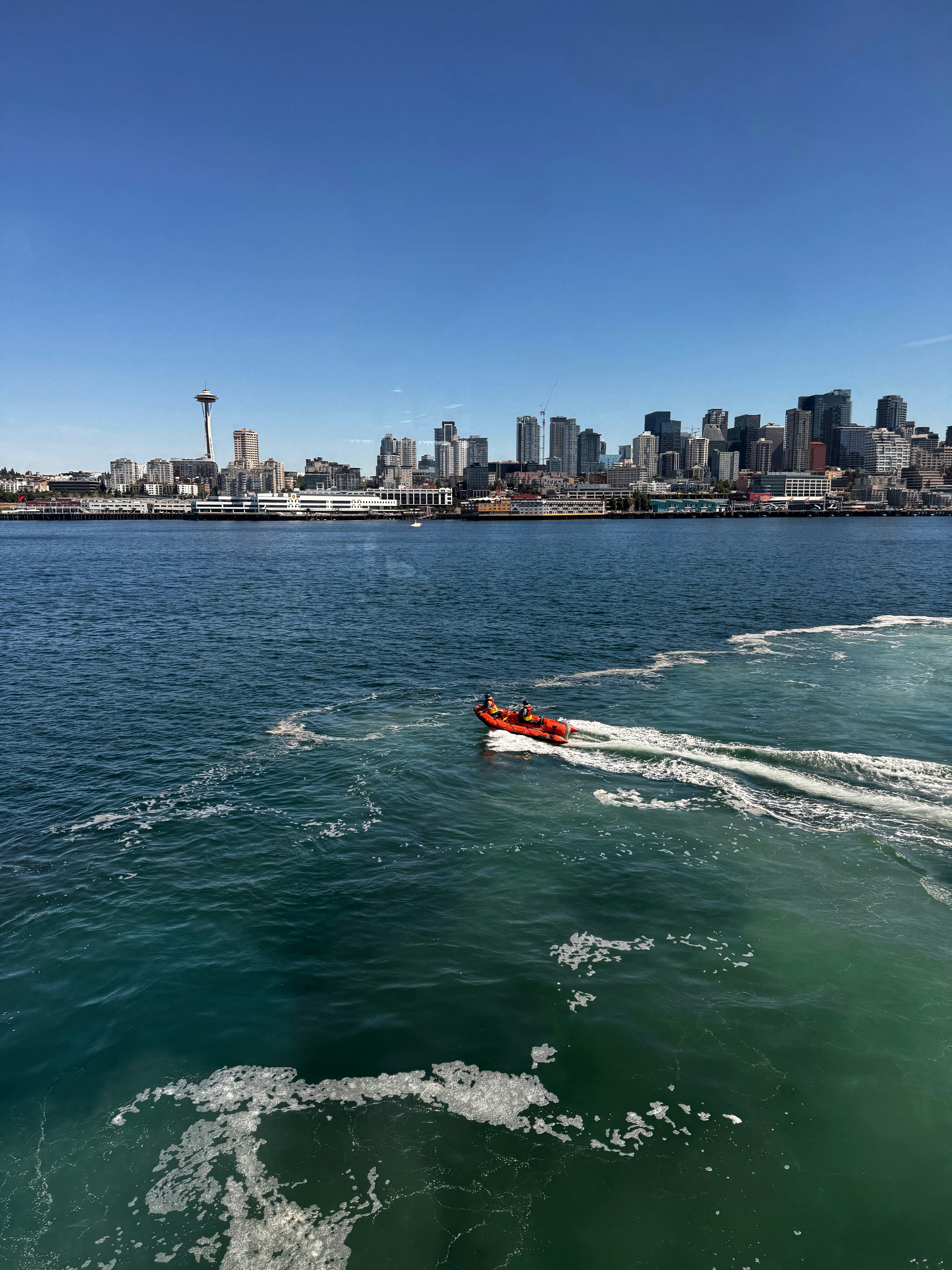 Orange boat speeds across blue water with city skyline.