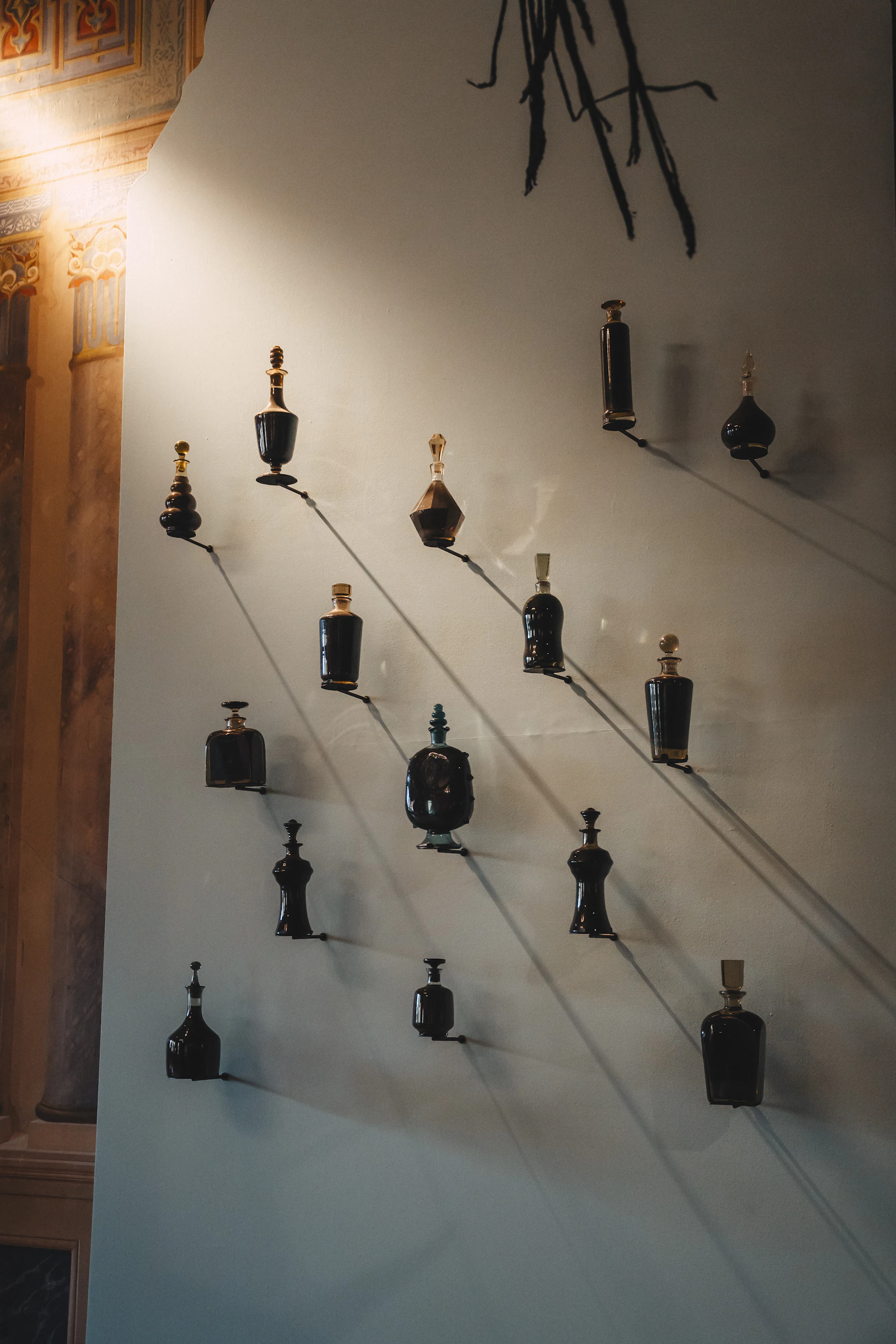Fourteen dark antique perfume bottles with stoppers, each on its own small bracket on a pale wall