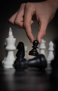 Hand moving a black pawn over fallen chess pieces.
