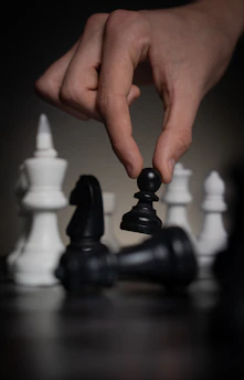 Hand moving a black pawn over fallen chess pieces.
