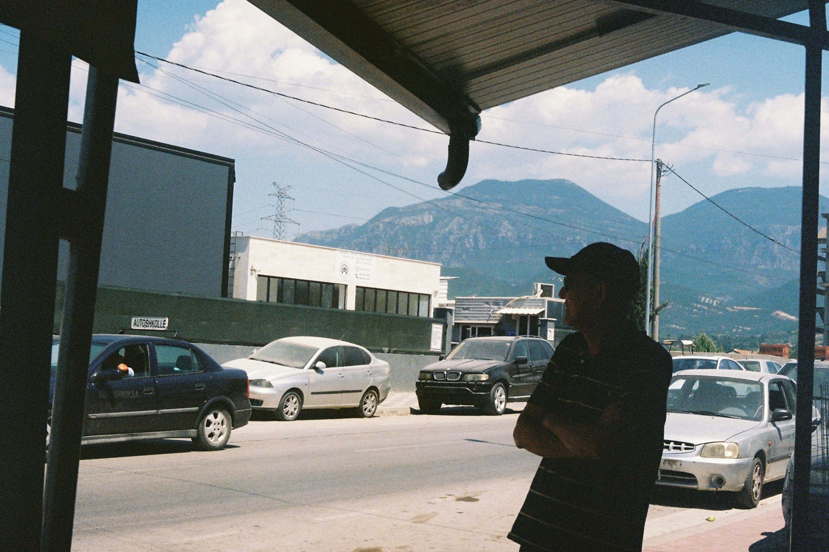 Man standing at bus stop with cars and mountains.