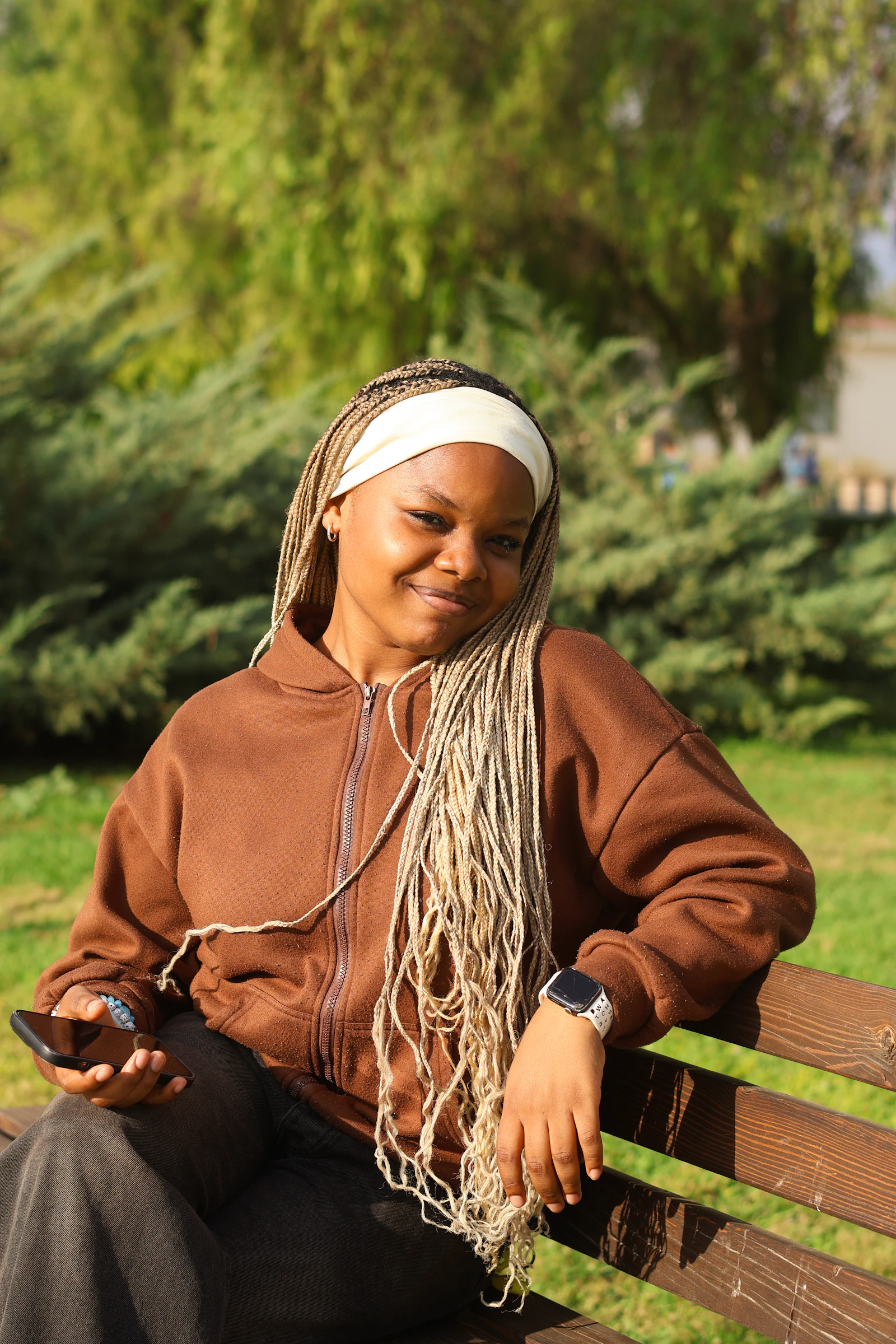 A smiling Black woman with long braids and a cozy brown sweatshirt sits on a park bench in the sun
