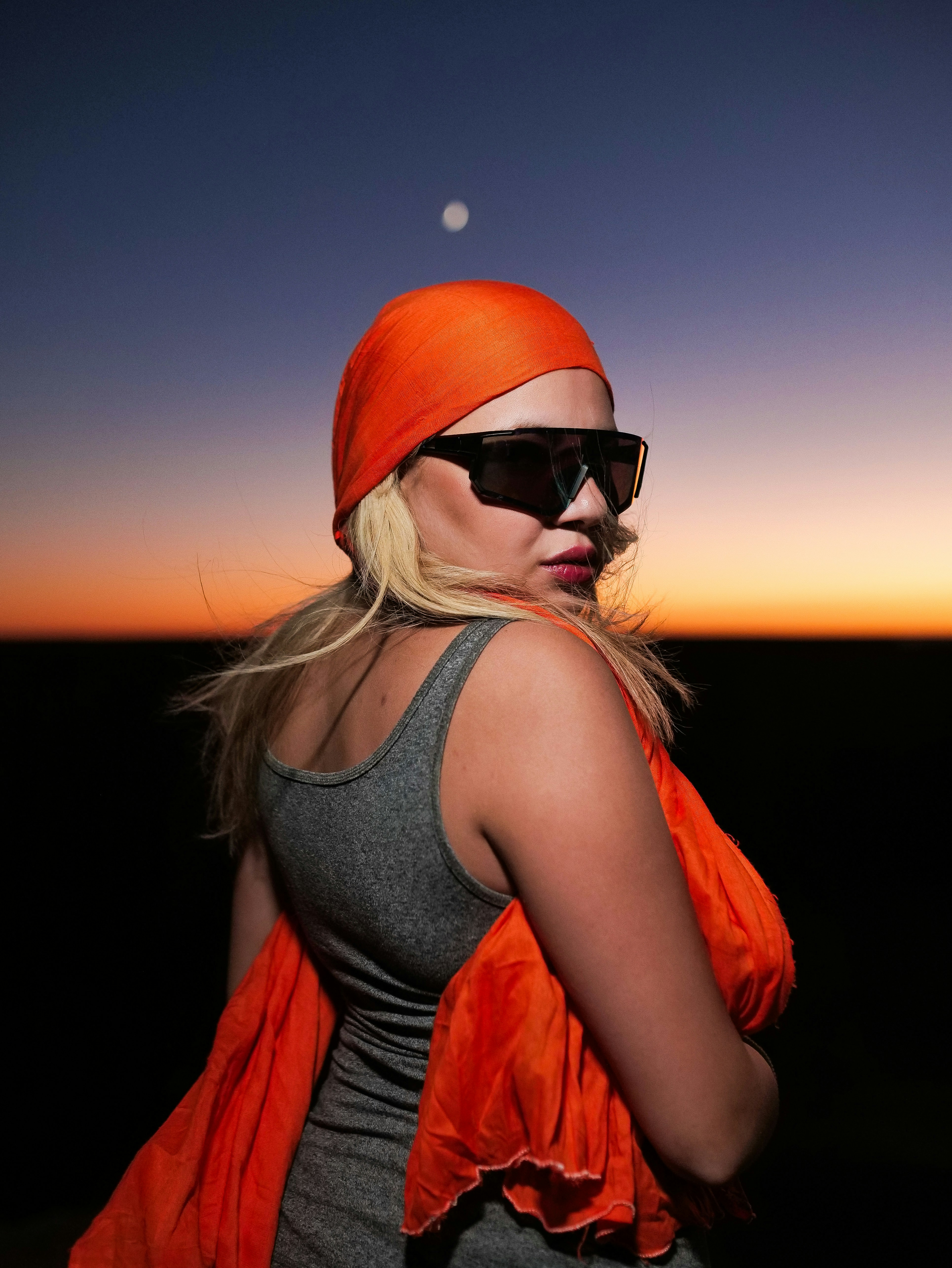 Woman in sunglasses and orange headscarf at sunset.