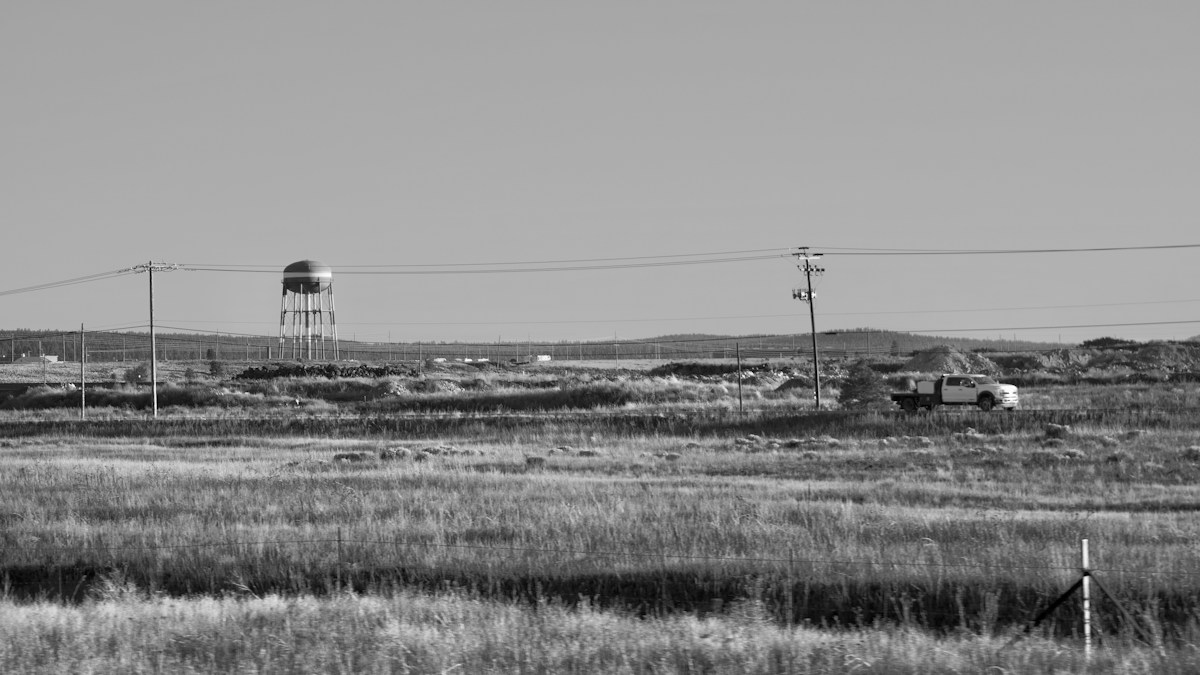 Château d’eau isolé au milieu d’une plaine d’herbes sèches, un pick-up sur la route, photo en noir et blanc