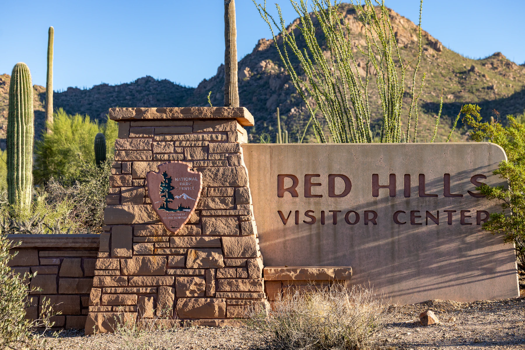 A stone sign reading Red Hills Visitor Center with the National Park Service arrowhead, among saguaro cacti