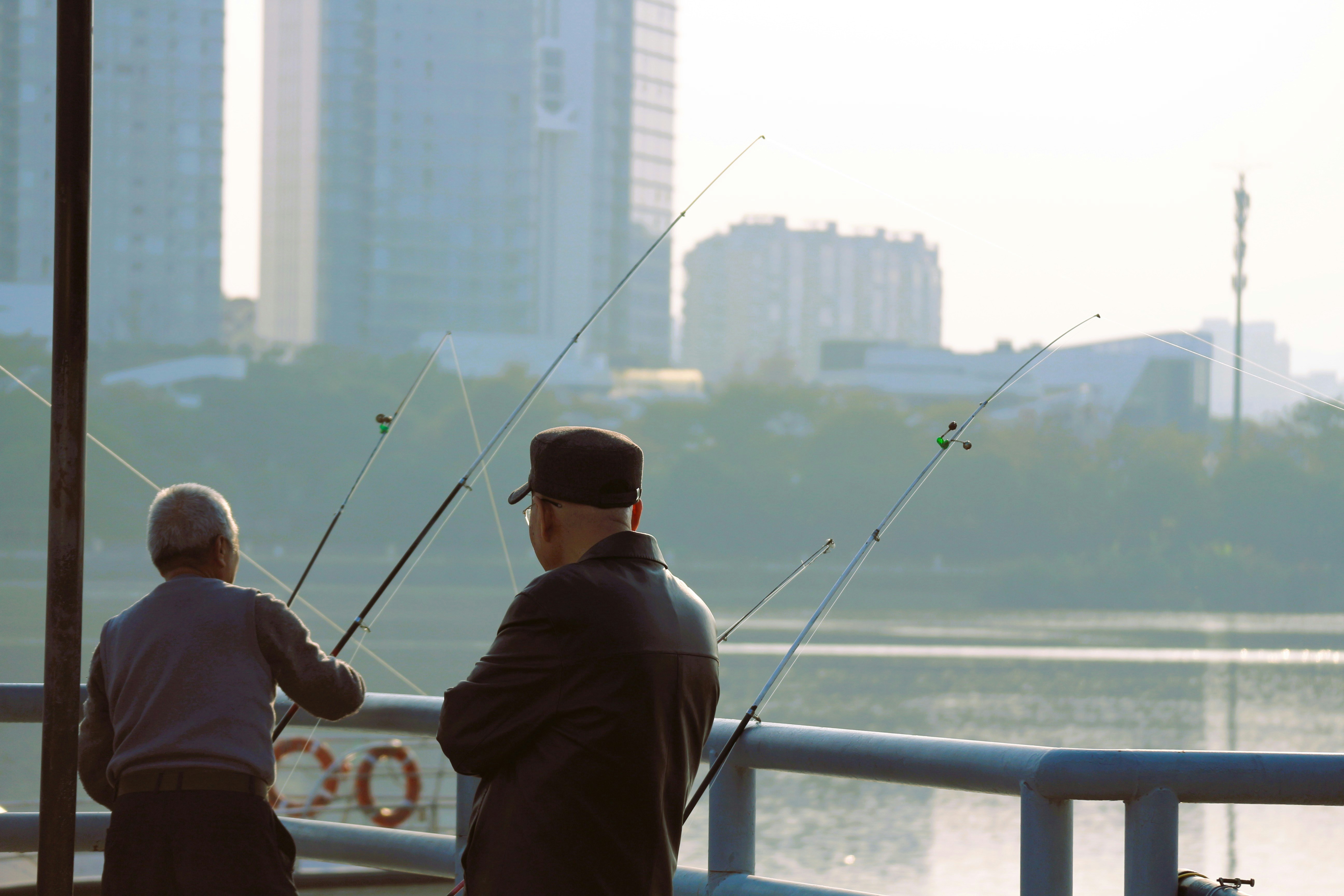 Two men fishing on a pier with city buildings.
