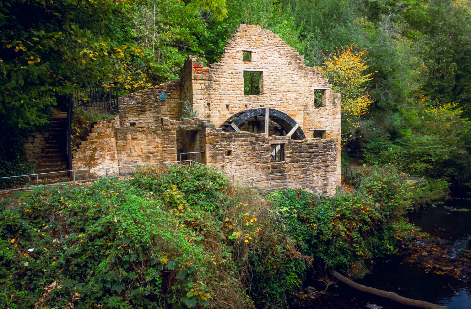 A ruined stone mill with a rusted wheel standing among trees