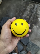 A hand holds a yellow smiley face stress ball.
