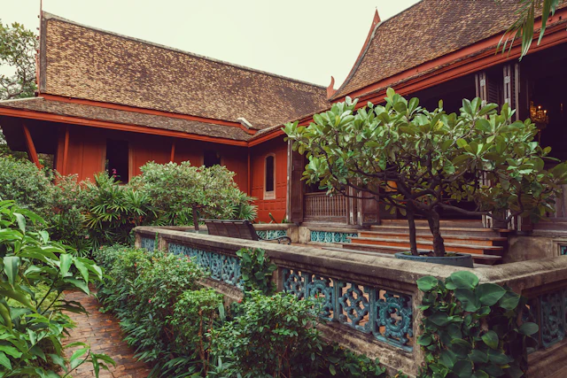 The red teak pavilions of Jim Thompson House seen across a lush tropical garden with a tiled balustrade.