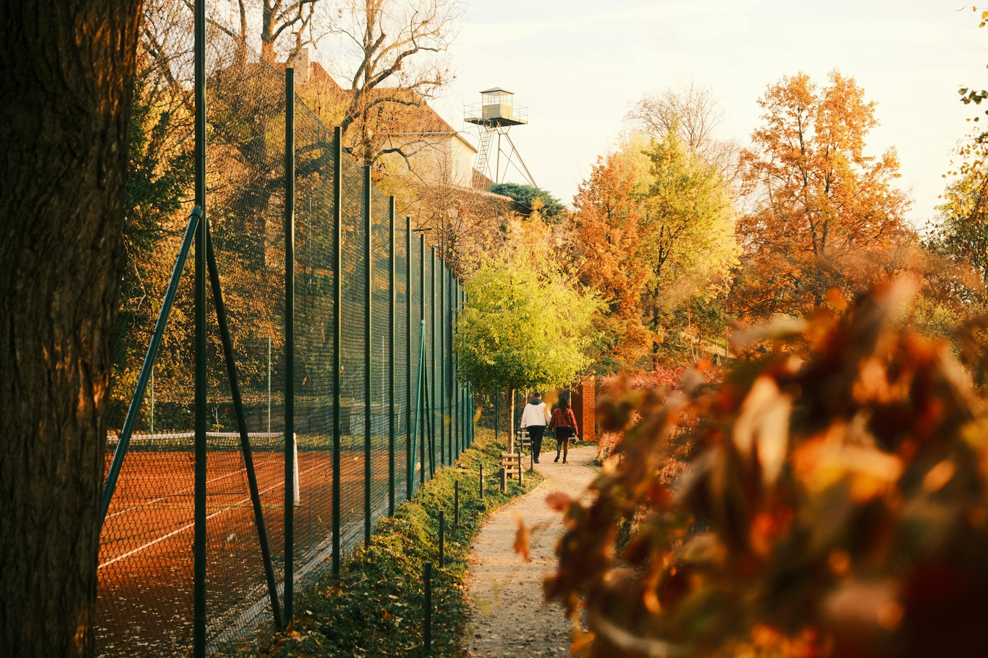 People walking on path beside tennis court in autumn.