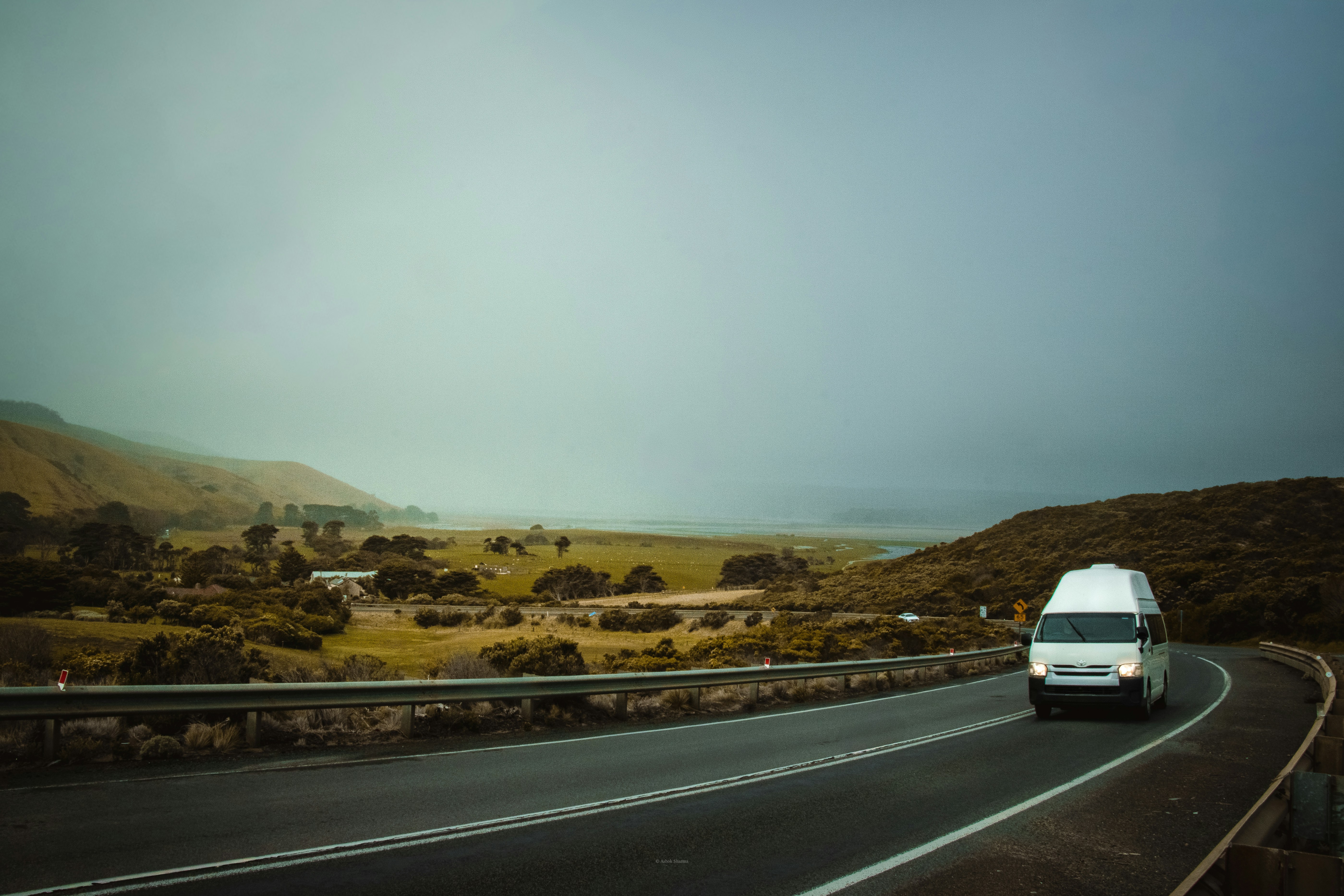 A white van drives on a highway along rolling coastal hills in Australia