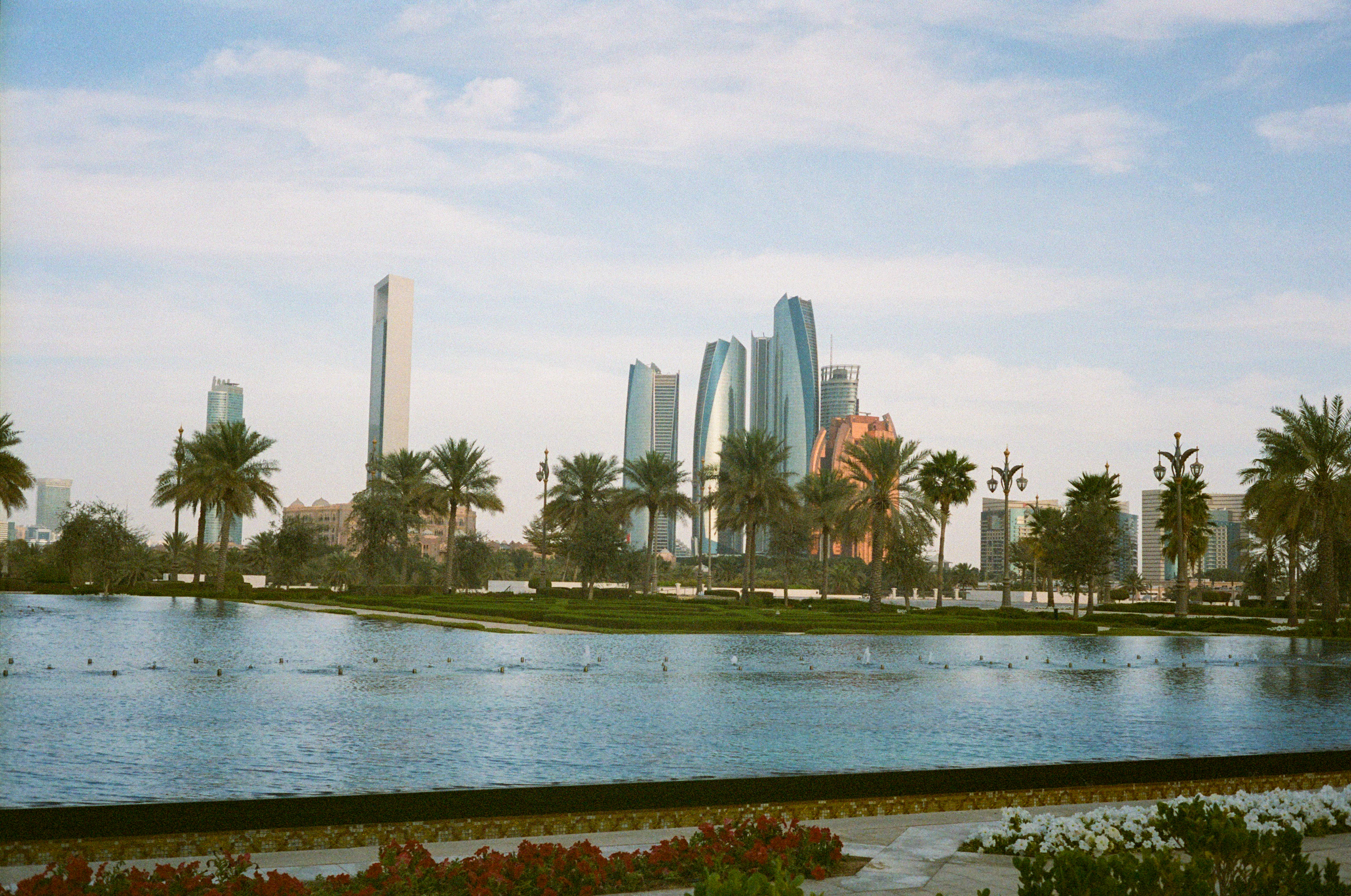 A green waterside park on the Abu Dhabi Corniche, lawns and date palms edging a still lagoon with birds on it, the Etihad Towers rising behind