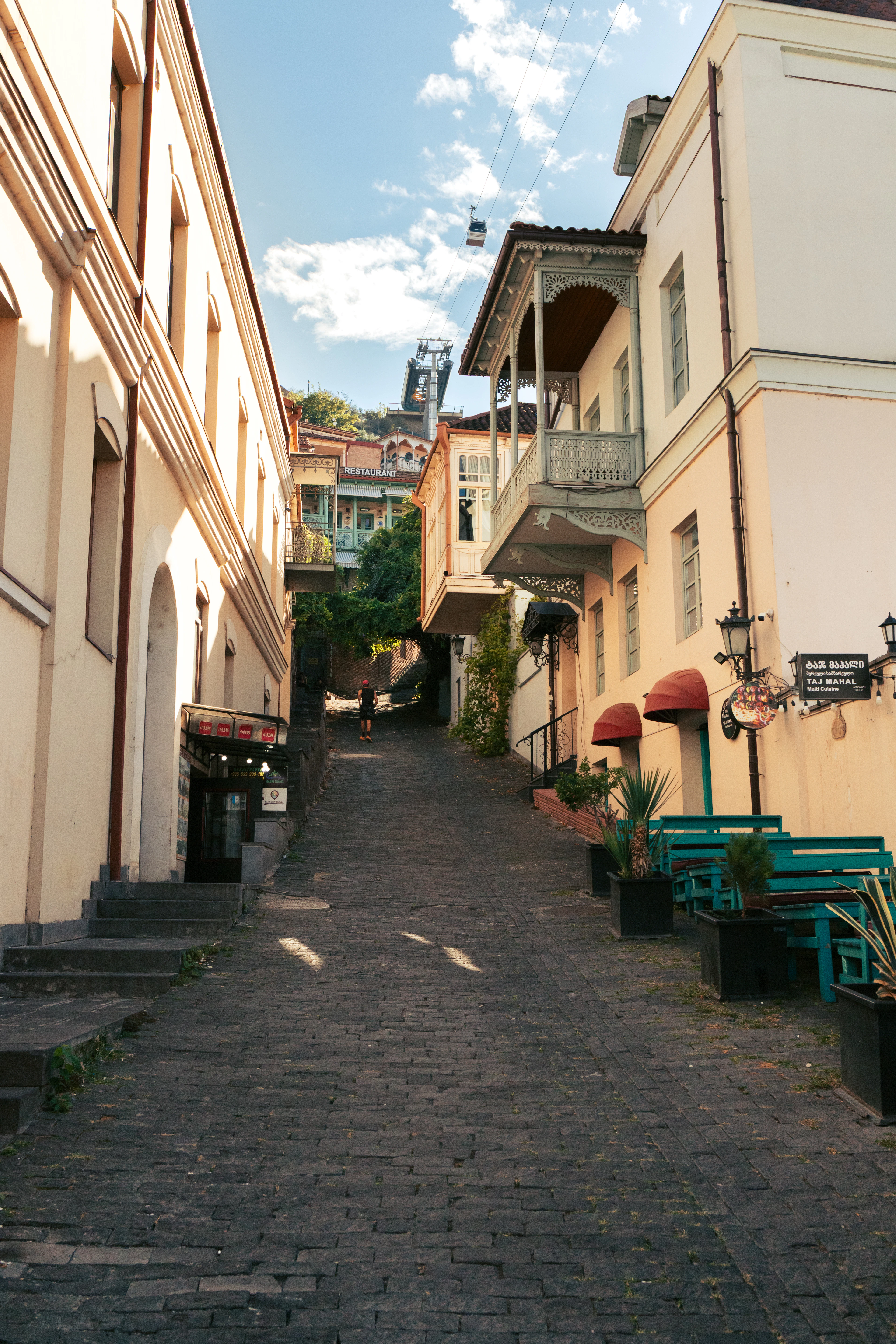 A cobbled backstreet climbing through Tbilisi's old town in warm morning light, a carved wooden balcony overhanging on the right and a cable car cabin crossing the sky towards Narikala above