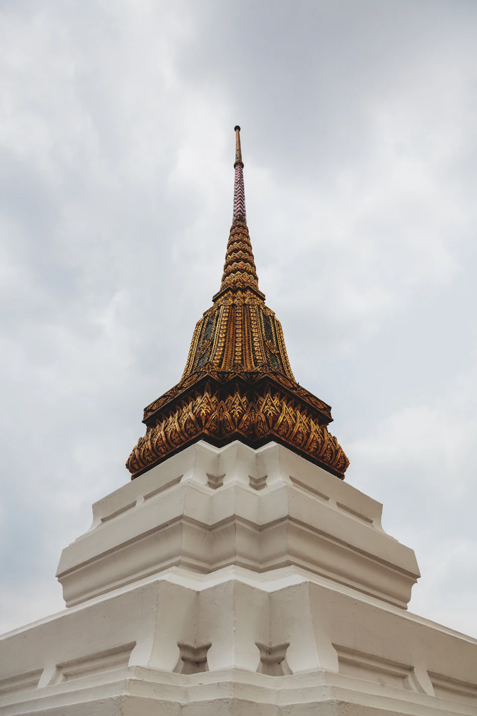 The image shows a majestic white temple with a golden spire reaching towards the sky, standing tall against an overcast backdrop. The temple is adorned with intricate gold designs and patterns on its surface, adding to its grandeur. The perspective of the photo is from below, looking up at the temple, emphasizing its height and prominence in the scene.

The image also contains some text that reads "สมายงทระนี้จำกลายง", which translates to "the sky is cloudy".