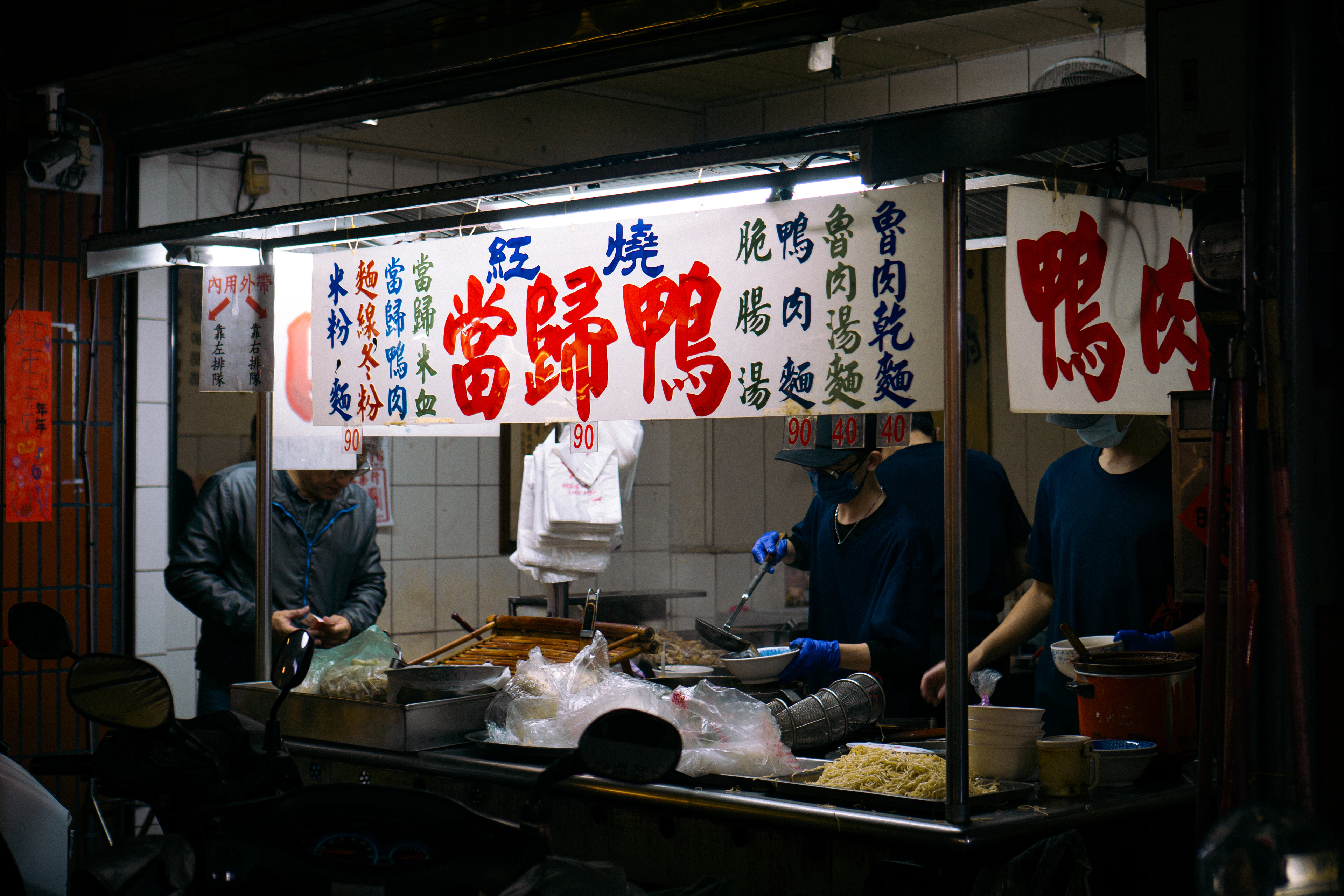 A Taiwanese street-food stall at night, its hand-painted boards in traditional Chinese advertising braised angelica duck and duck noodle soup, cooks working behind the counter.