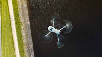 A white drone flies over dark water.