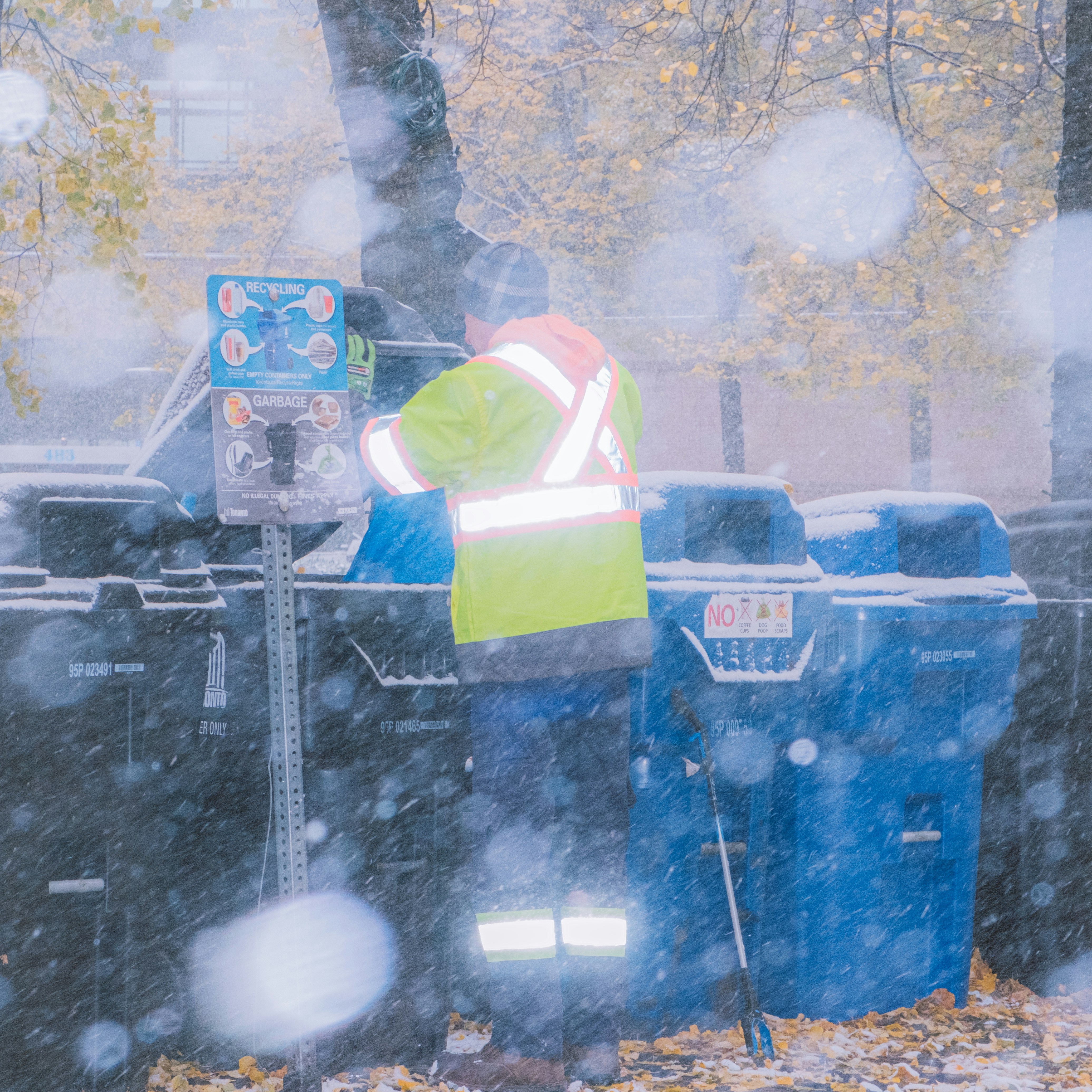 Worker in reflective vest near trash bins in snow.