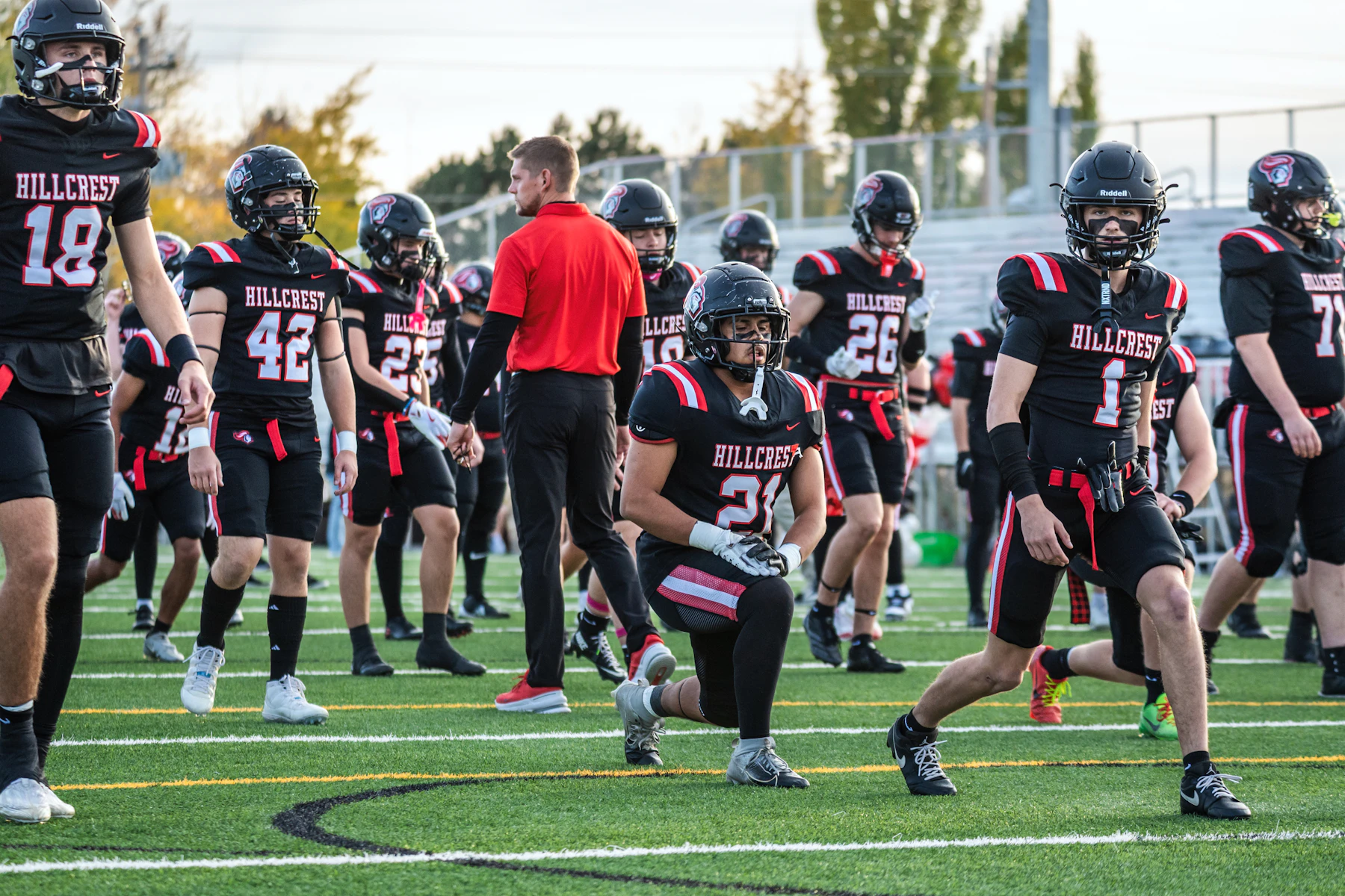 A football team in black and red jerseys and helmets warming up on a turf field beside a coach