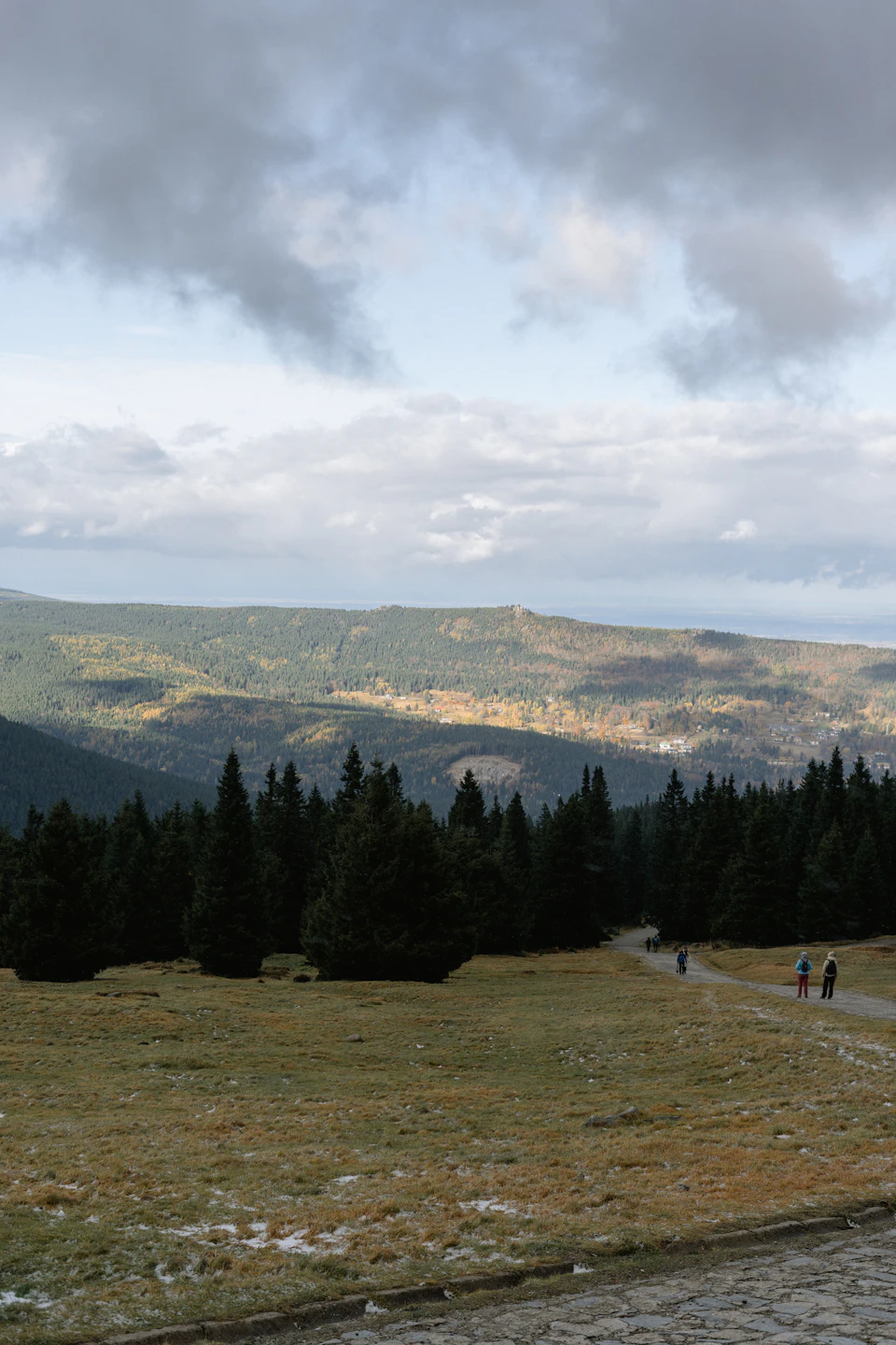 The image depicts a group of people walking along a dirt path that winds through a lush, green forested area. The sky above is filled with clouds, casting a soft light over the scene and creating an atmosphere of tranquility. The perspective of the photo suggests it was taken from a higher vantage point, providing a sweeping view of the landscape below.