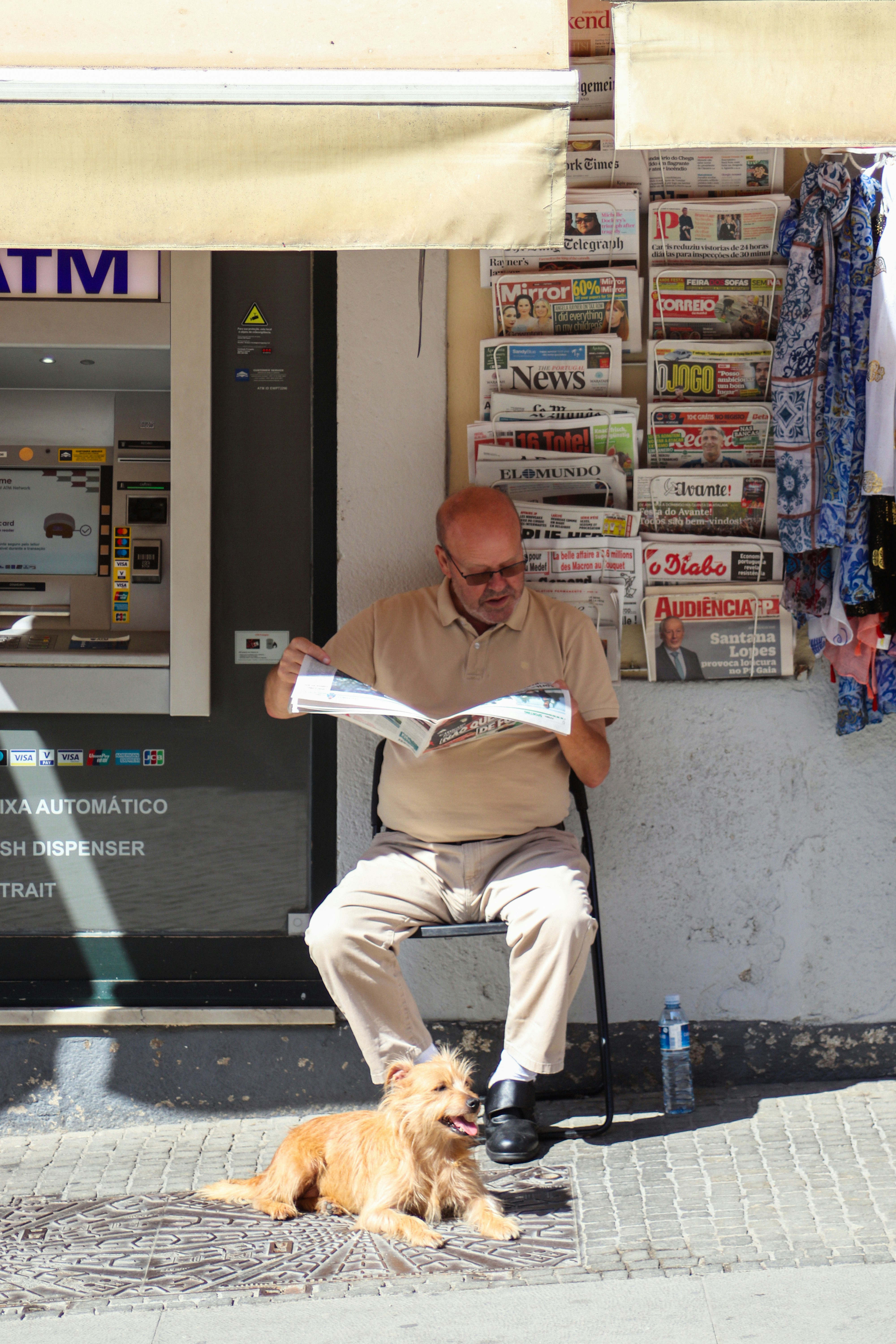 Man reading newspaper with dog beside atm.