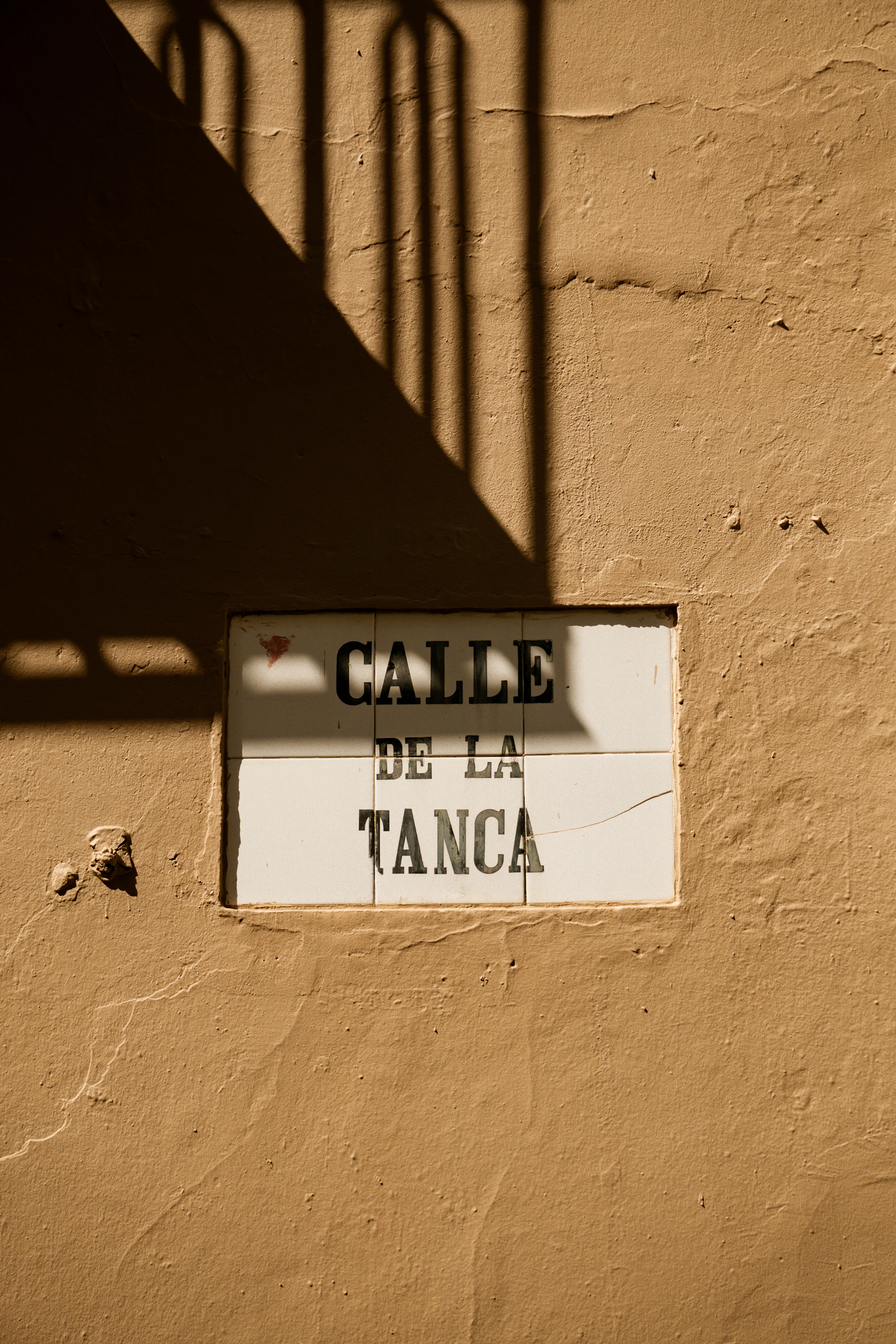 A white tile street plaque reading Calle de la Tanca on an ochre wall, crossed by the shadow of a balcony