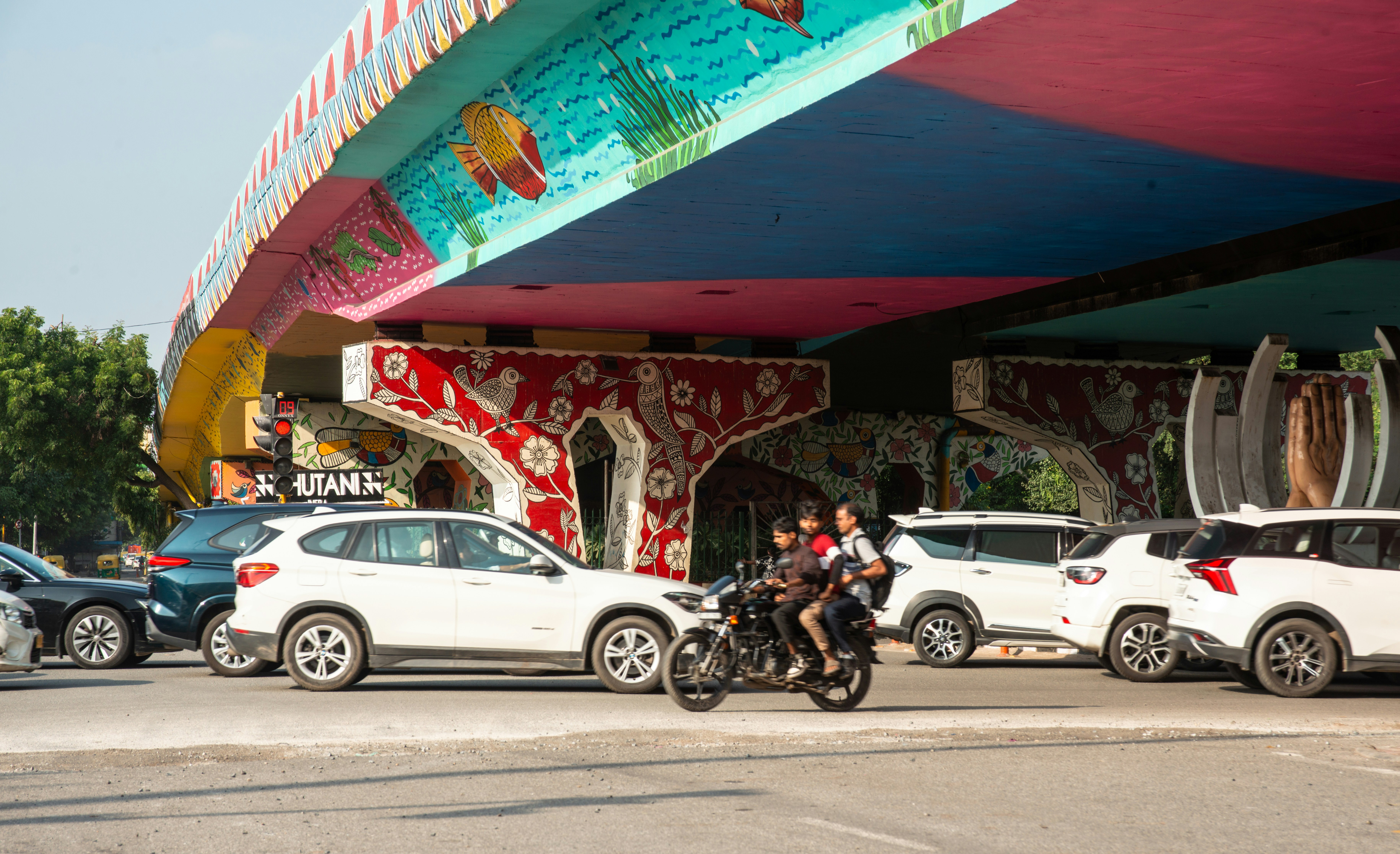 A colorful painted flyover in Delhi, India, featuring vibrant Madhubani-style art under the bridge with cars passing below. The mural showcases fish, birds, and floral motifs in red, blue, yellow, and green, reflecting India’s street art culture and efforts to beautify public spaces.