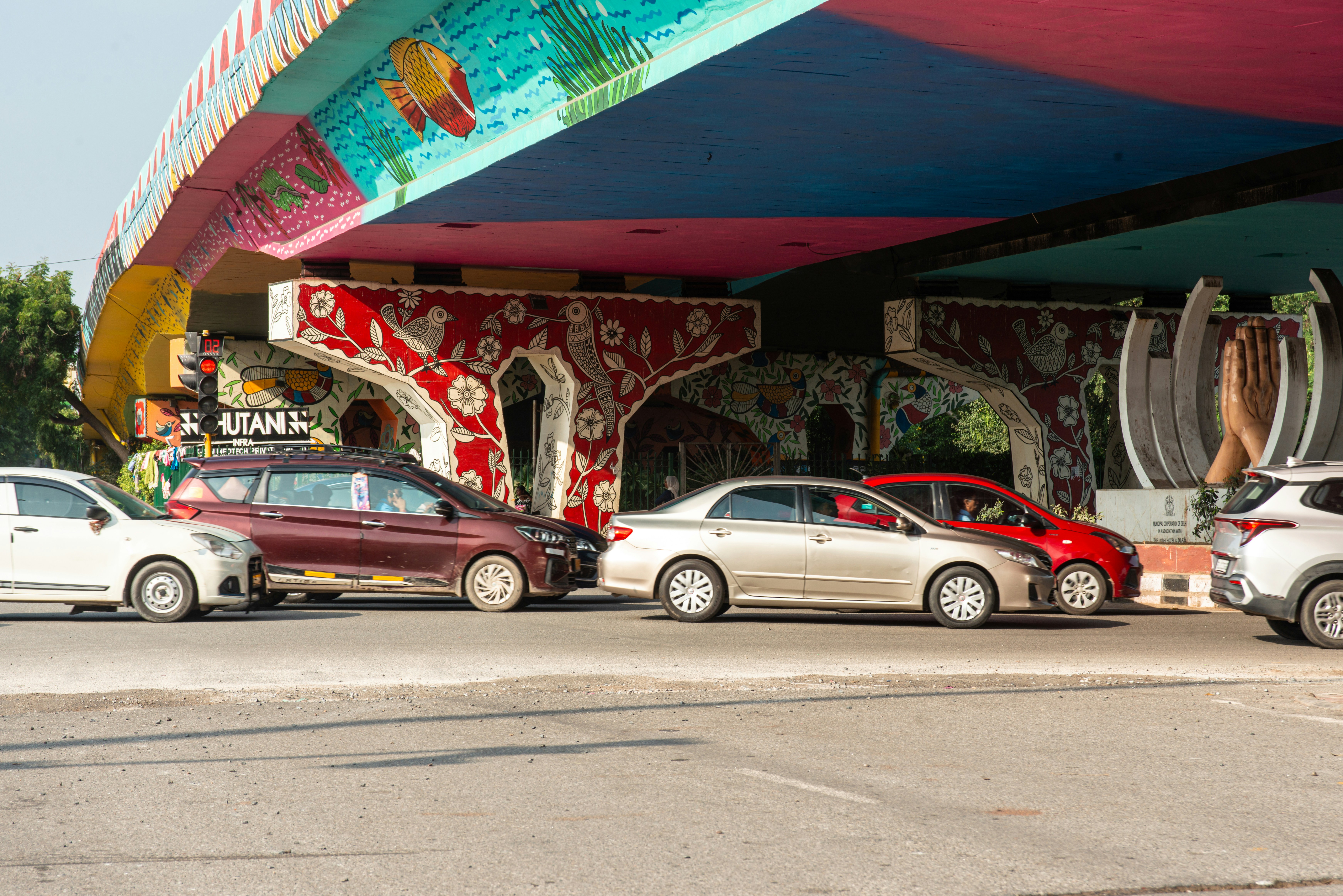 A colorful painted flyover in Delhi, India, featuring vibrant Madhubani-style art under the bridge with cars passing below. The mural showcases fish, birds, and floral motifs in red, blue, yellow, and green, reflecting India’s street art culture and efforts to beautify public spaces.
