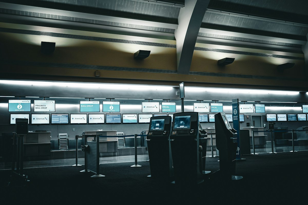 An empty airport check-in hall, a row of dark self-service kiosks in the centre and behind them empty counters and a row of screens glowing pale blue
