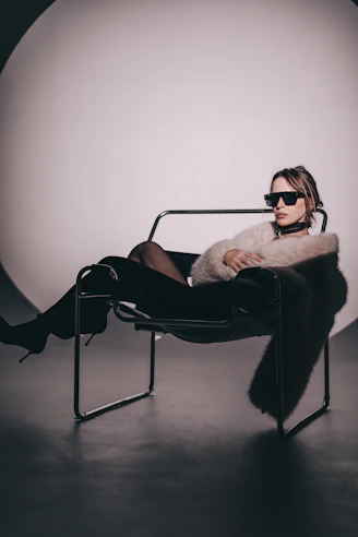 Woman in sunglasses and fur jacket sits in chair.