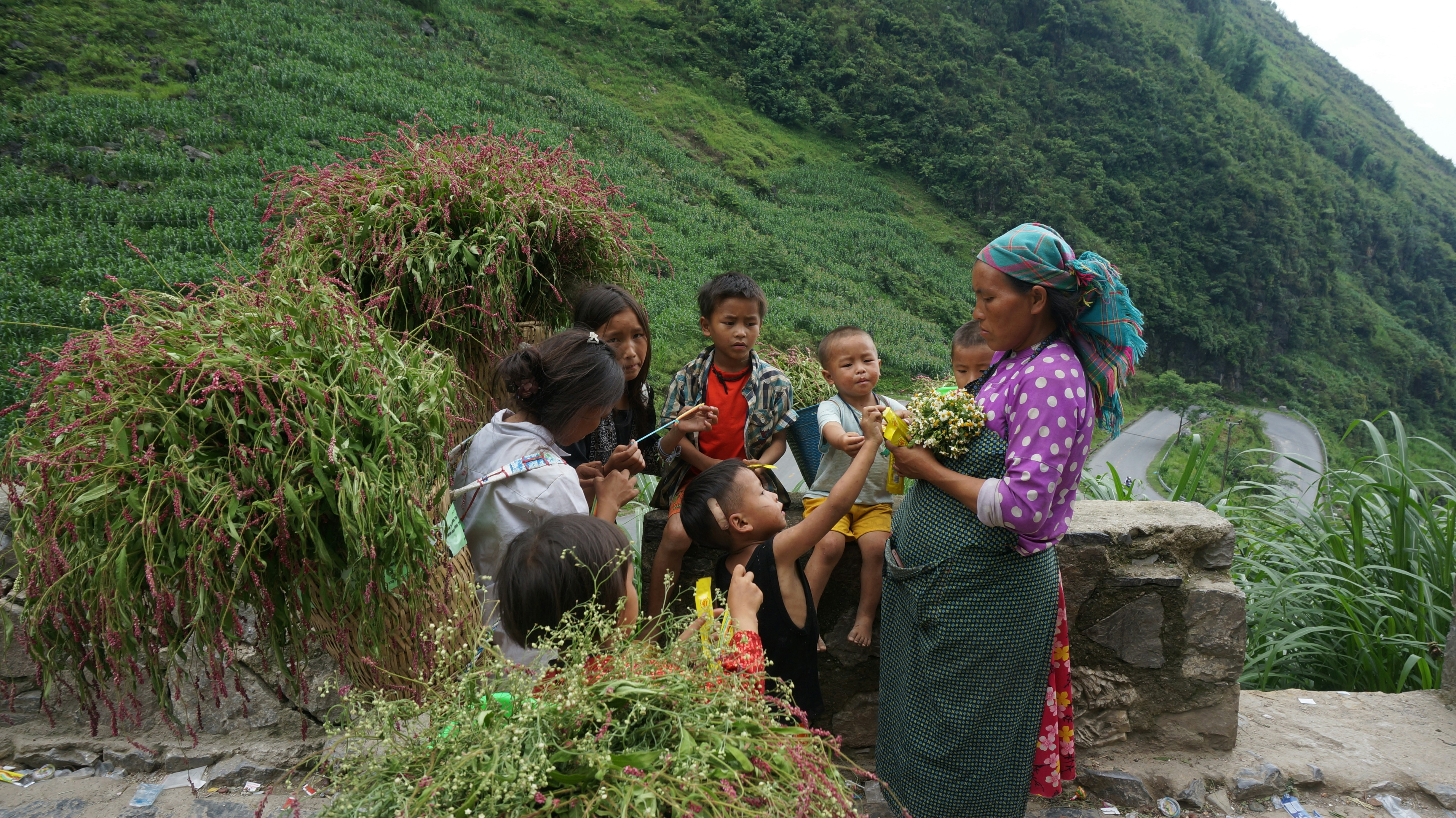 A chaotic scene showing the aftermath of a flash flood or landslide in a mountain village. Debris, damaged houses, and a muddy river overflowing its banks are visible, with rescue workers in the background.