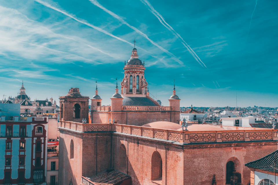 The image presents an aerial view of a cityscape, dominated by a large, ornate brick building with a dome and multiple towers. The building is situated on a hillside, providing a unique perspective of the surrounding area. In the distance, other buildings can be seen, contributing to the urban landscape. The sky above is a clear blue, dotted with wispy white clouds that add depth to the scene.