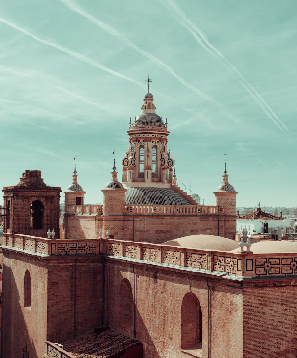 The image depicts a majestic brick building with a dome and multiple towers, situated atop a hill. The building features intricate carvings on its facade, adding to its architectural beauty. The perspective of the photo is from below, looking up at the structure, emphasizing its grandeur. In the distance, there are other buildings visible in the background, suggesting that this particular building may be part of a larger cityscape or historic district.

The sky above the scene is clear and blue, with wispy clouds scattered across it. The image does not contain any discernible text or human figures, making the viewer focus solely on the architectural marvel before them.
