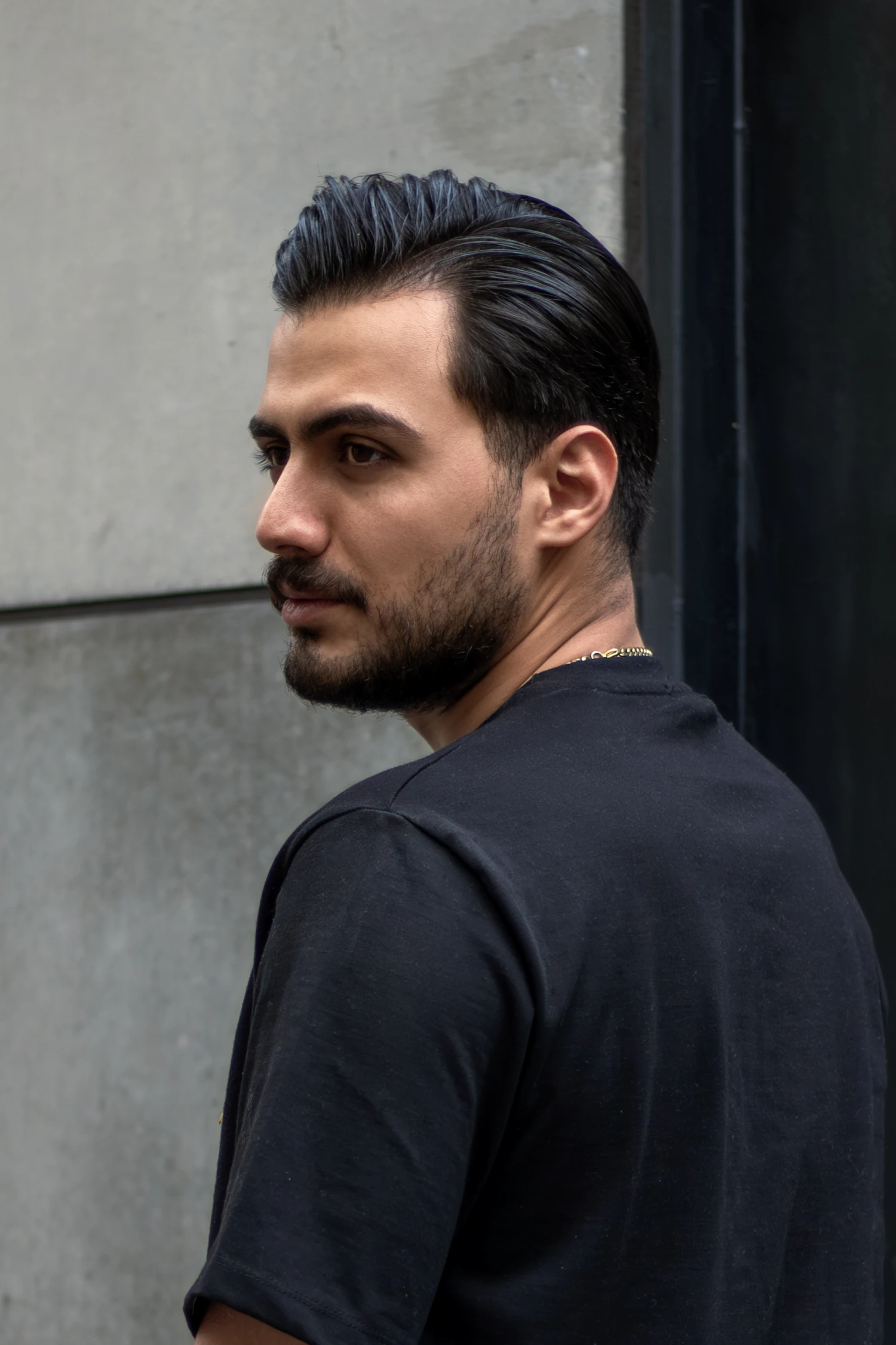 A man with slicked-back hair and a short beard standing against a grey wall